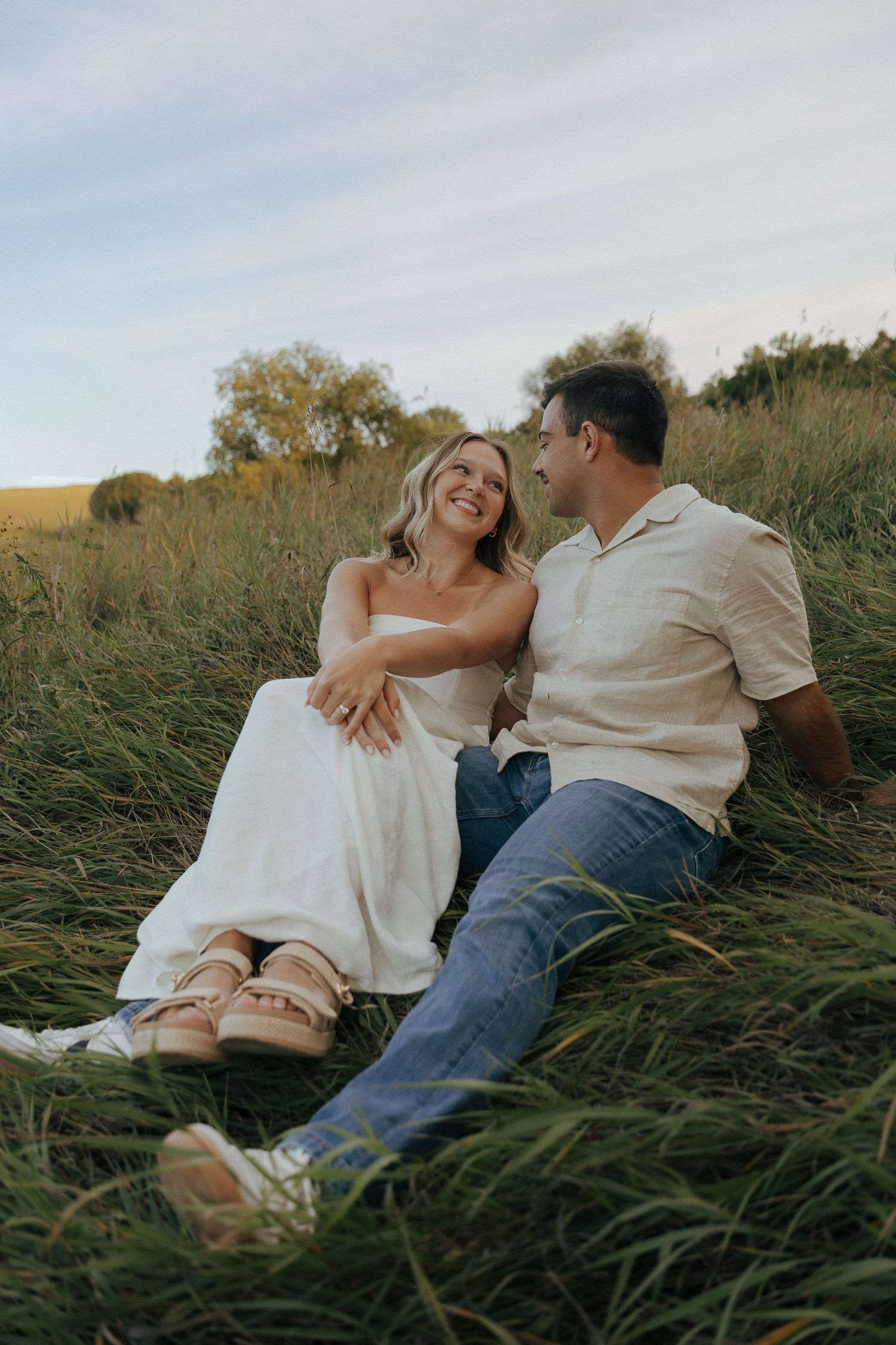 A couple relaxes together in a grassy field during a casual outdoor photoshoot.