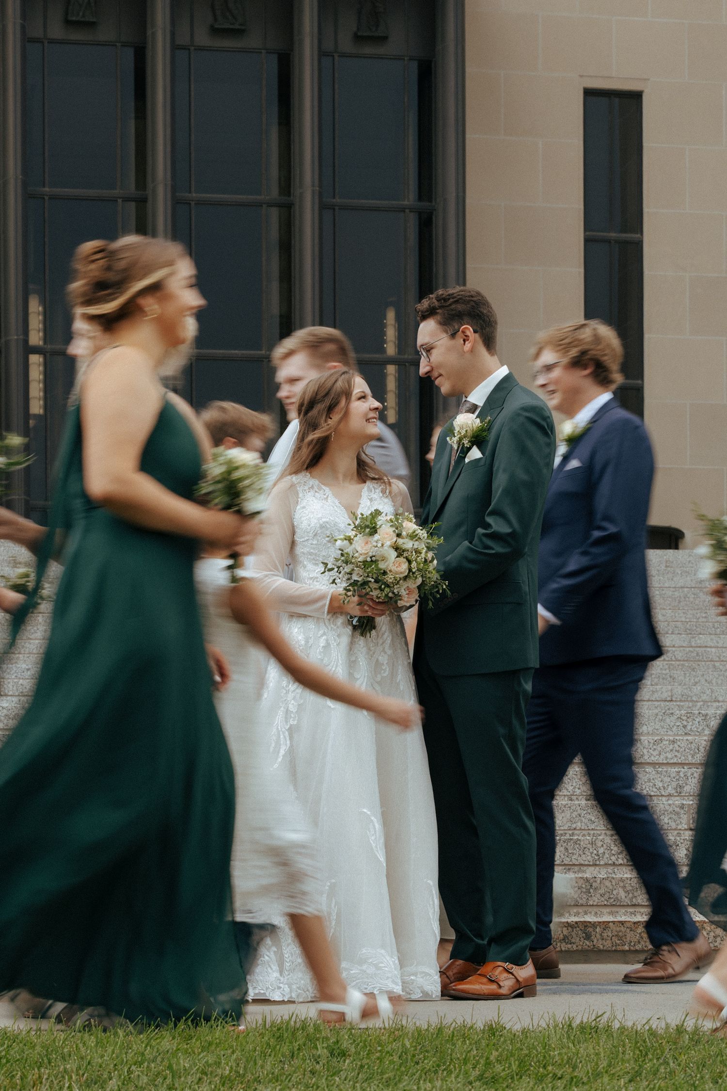 Wedding party members in green dresses and dark suits walk together outside a light-colored building.