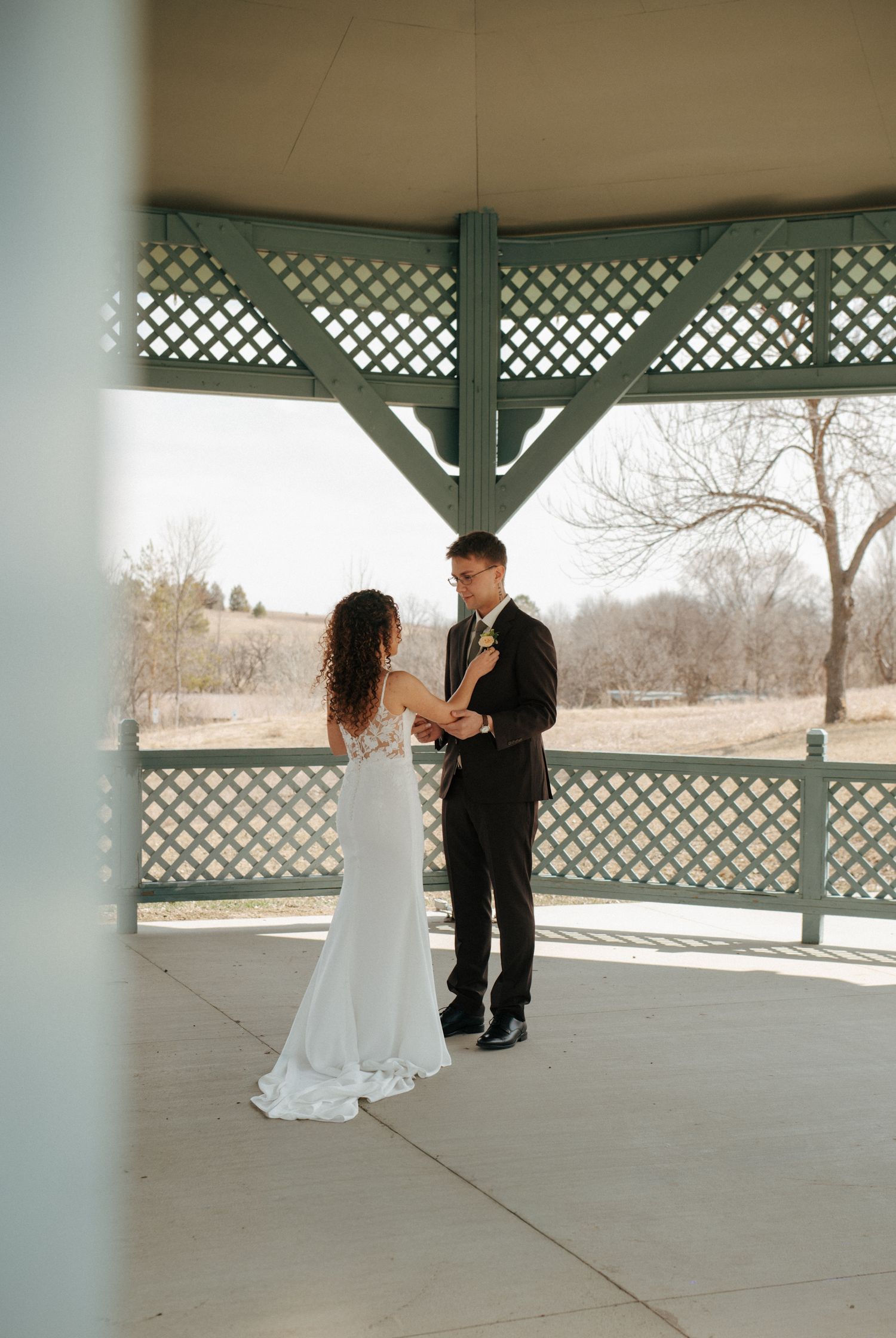 Couple exchanging vows in a green lattice gazebo, bride in white gown and groom in black tuxedo, outdoor winter wedding.