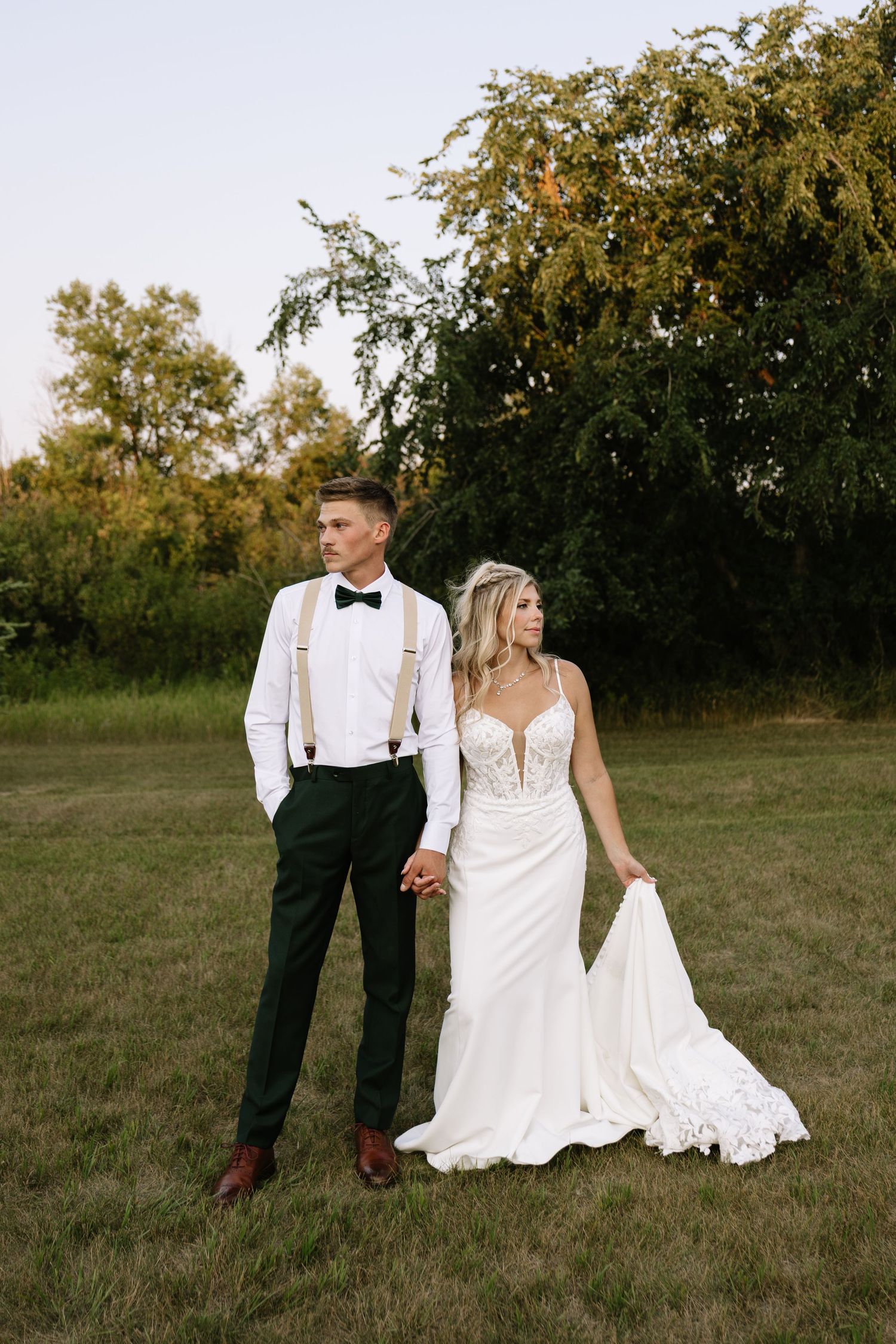 Bride and groom holding hands in a green field, groom in suspenders and bow tie, bride in lace wedding gown.