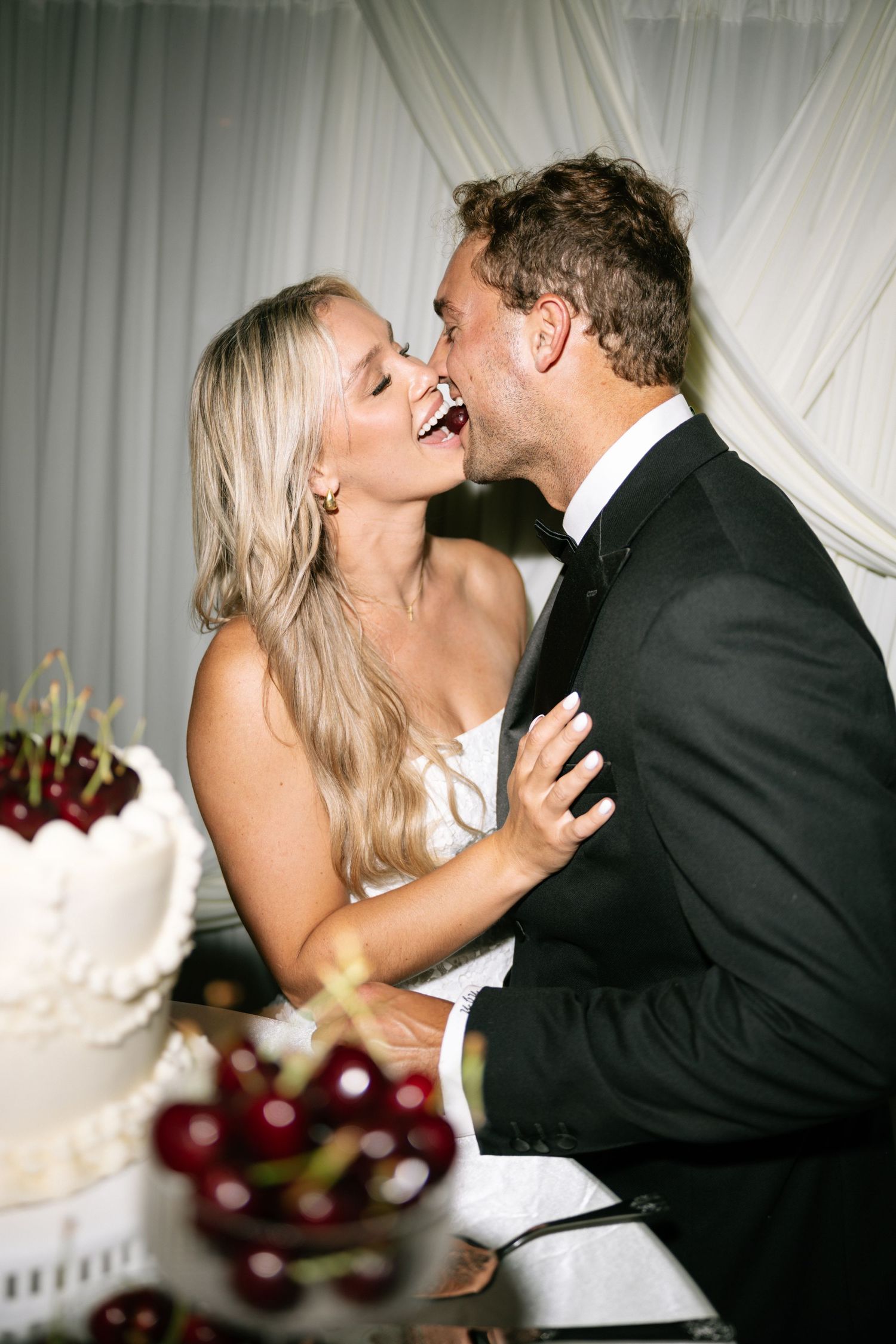 Bride and groom laughing and sharing a kiss beside a white wedding cake with cherries at their reception.