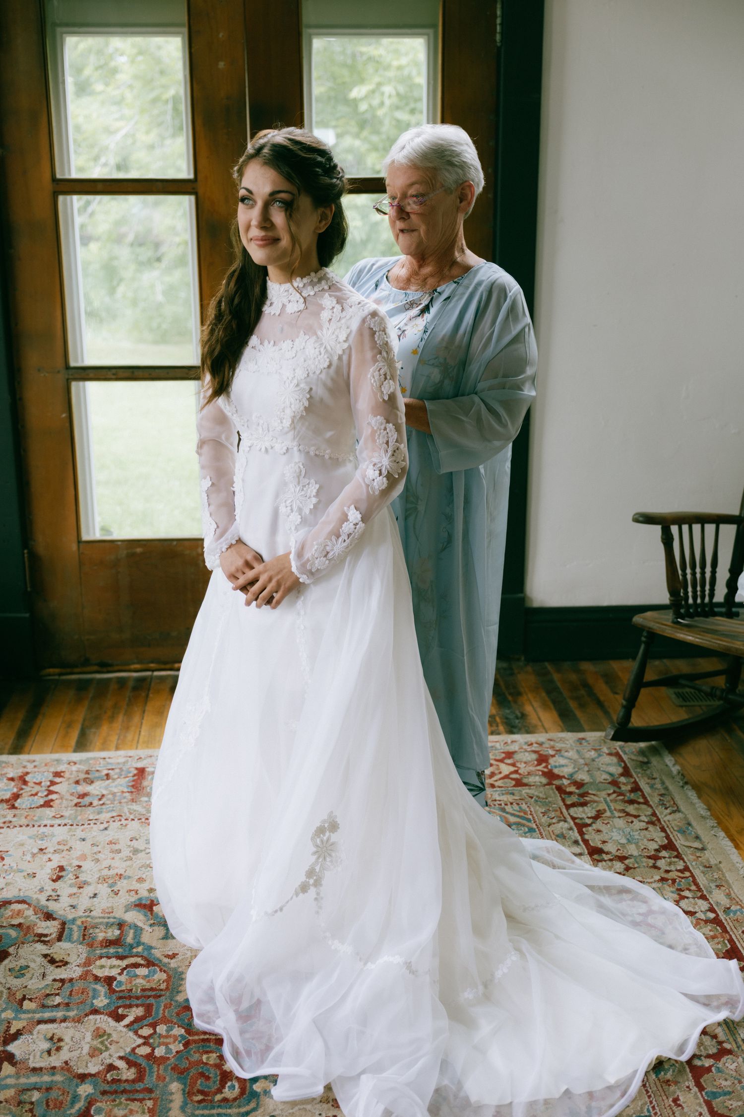 Bride in long-sleeve lace wedding gown being helped by older woman in blue dress indoors on patterned rug.