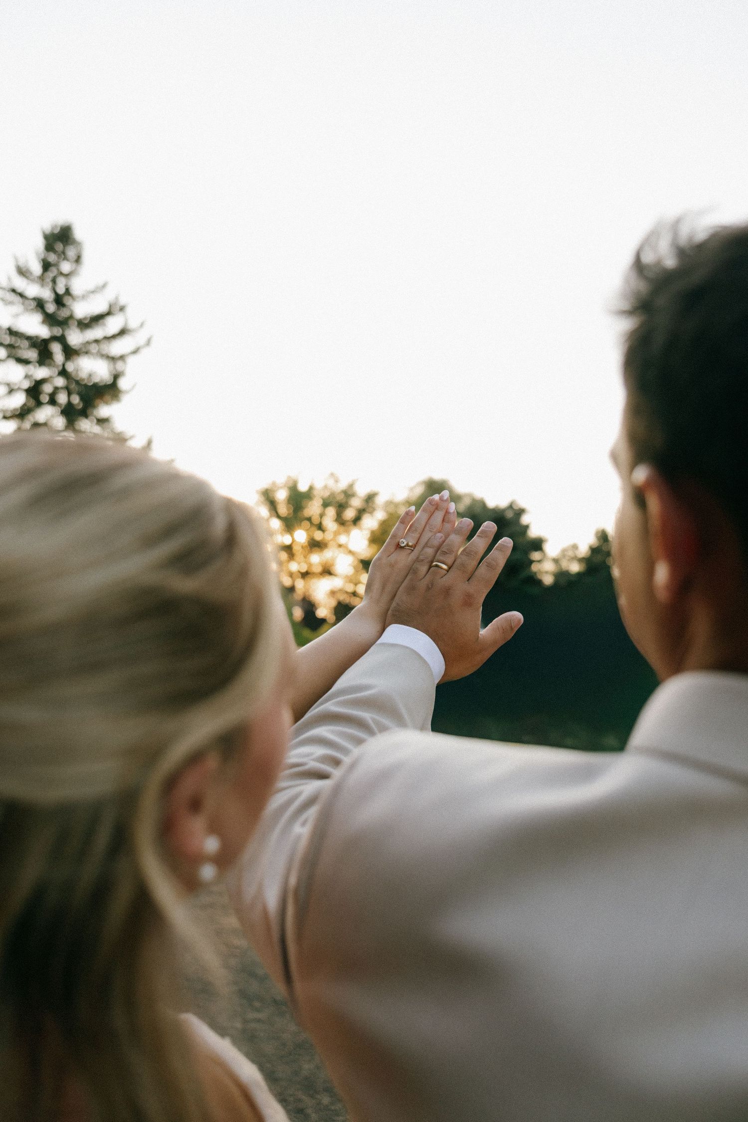 Newlywed couple holding hands showing wedding rings at sunset outdoors.