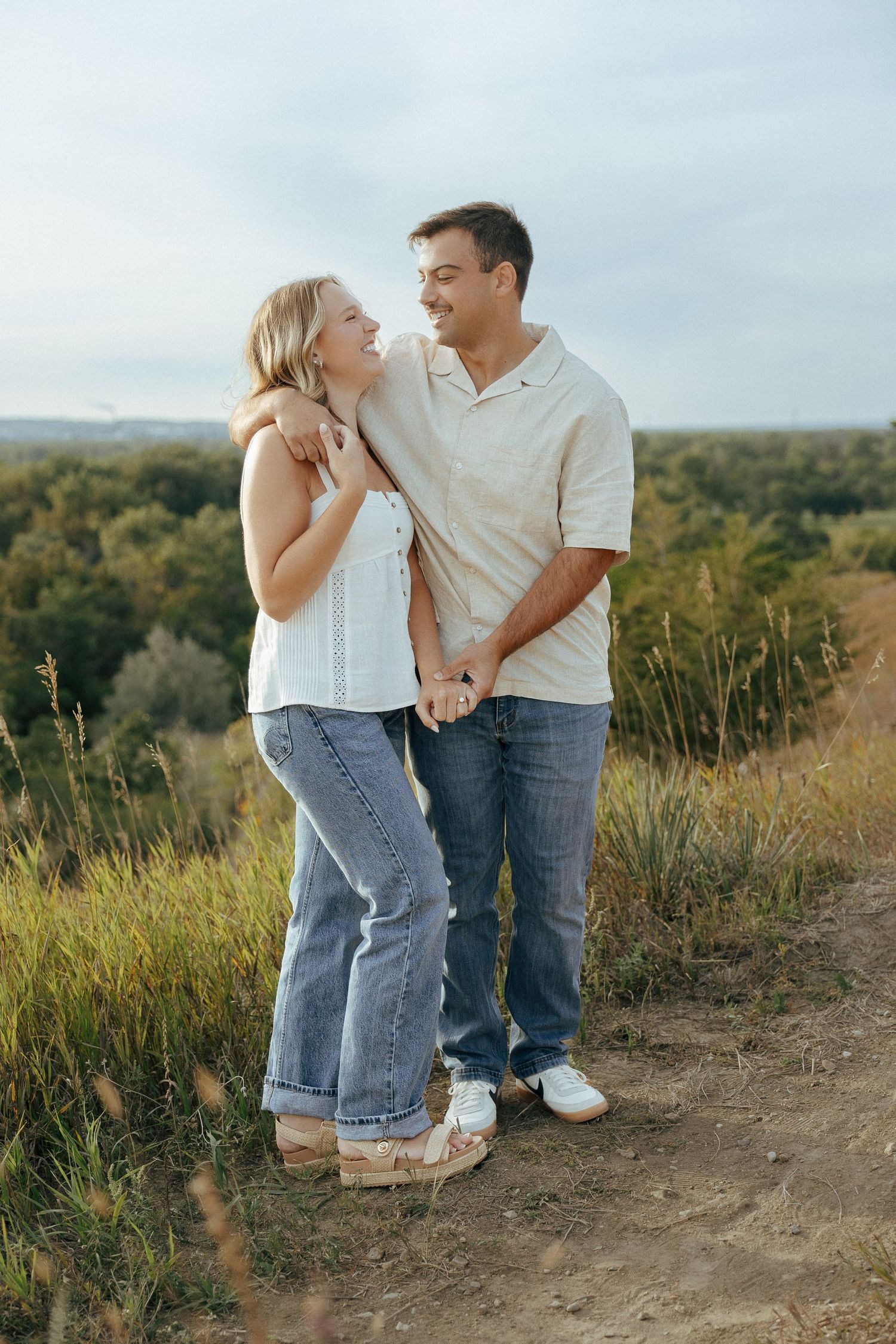 Two people in casual attire stand close together on a nature path with rolling hills in the background.