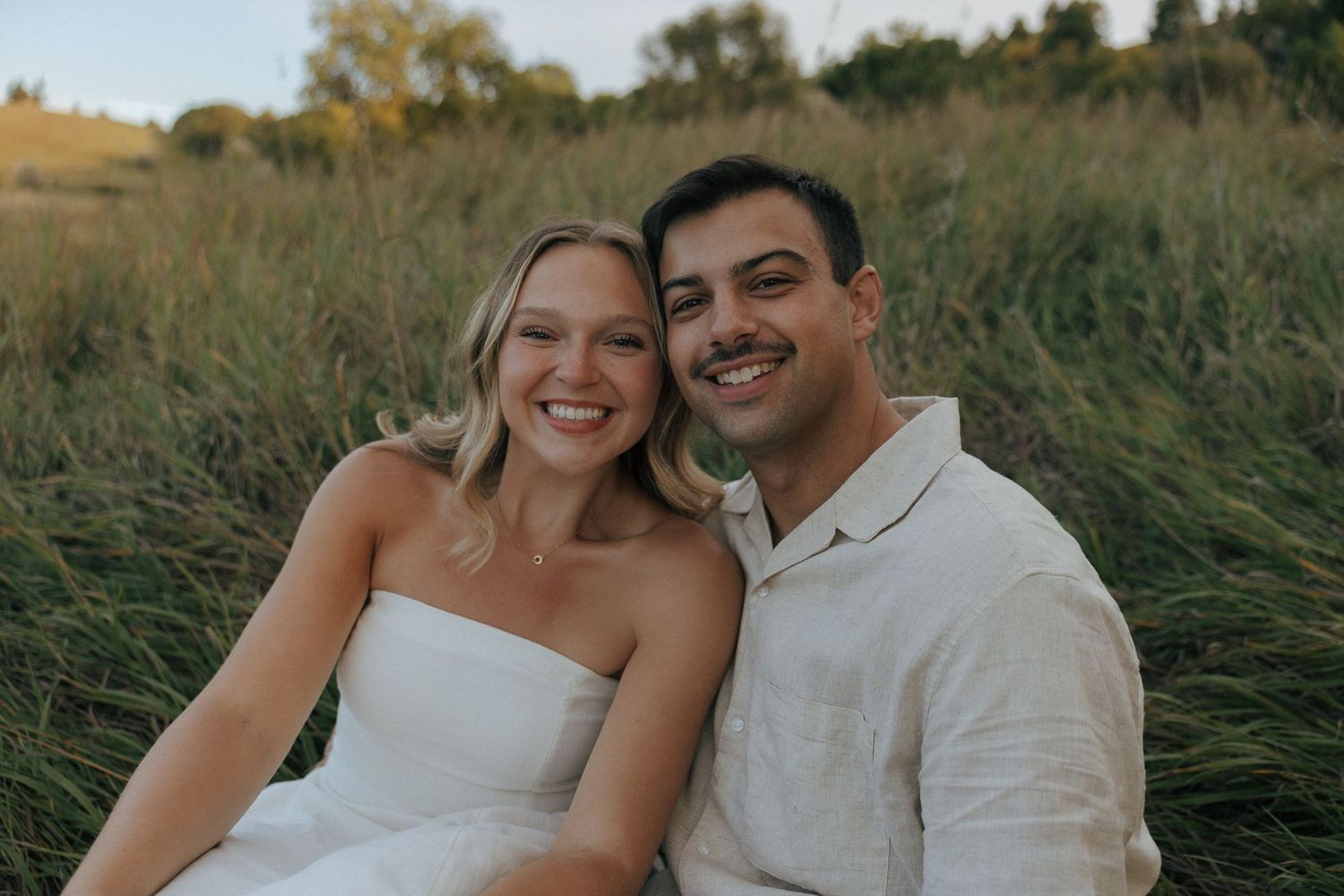 A couple poses for engagement photos in a scenic field with golden sunlight.