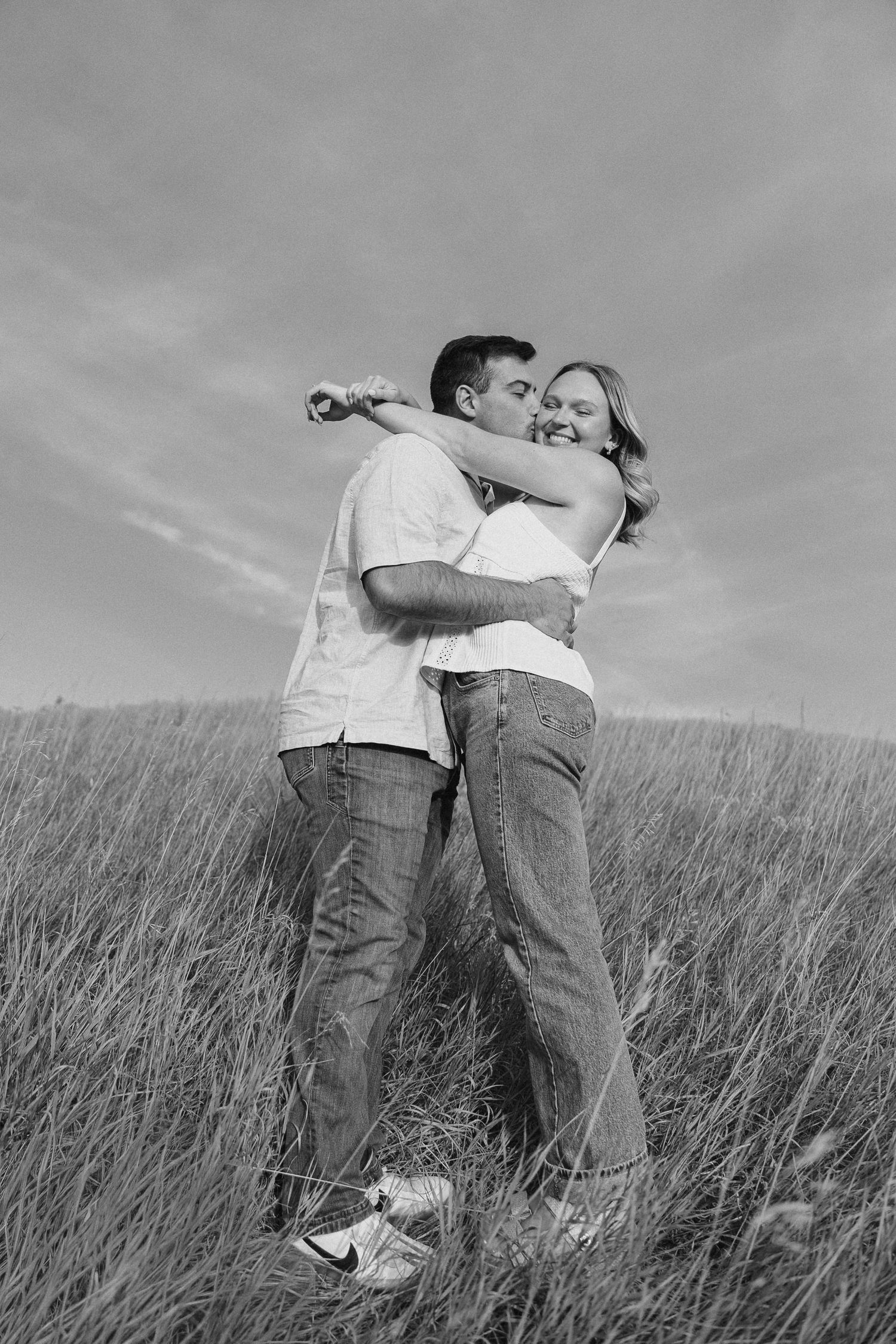 A black and white photo of two people embracing in a field of tall grass.