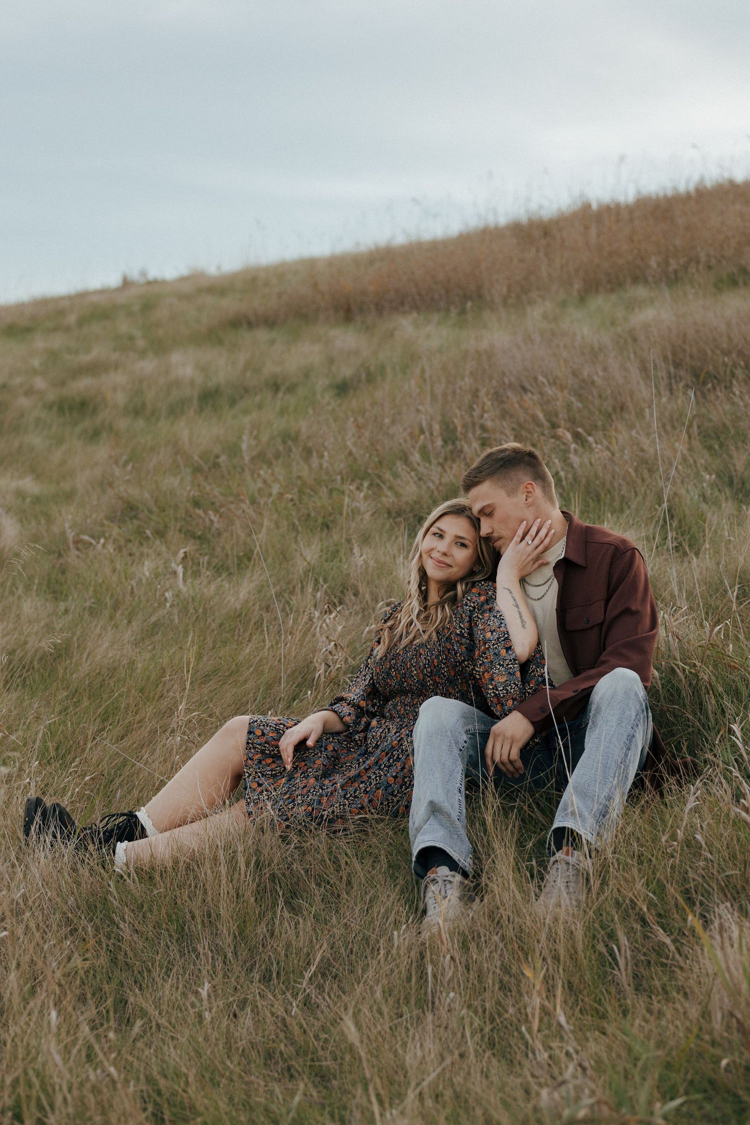 Romantic outdoor portrait of couple embracing on grassy hill under cloudy sky.