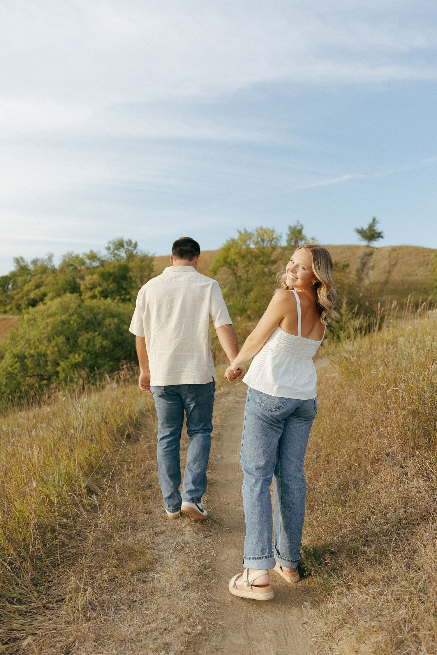 Two people stroll hand in hand along a winding dirt trail through golden grass.