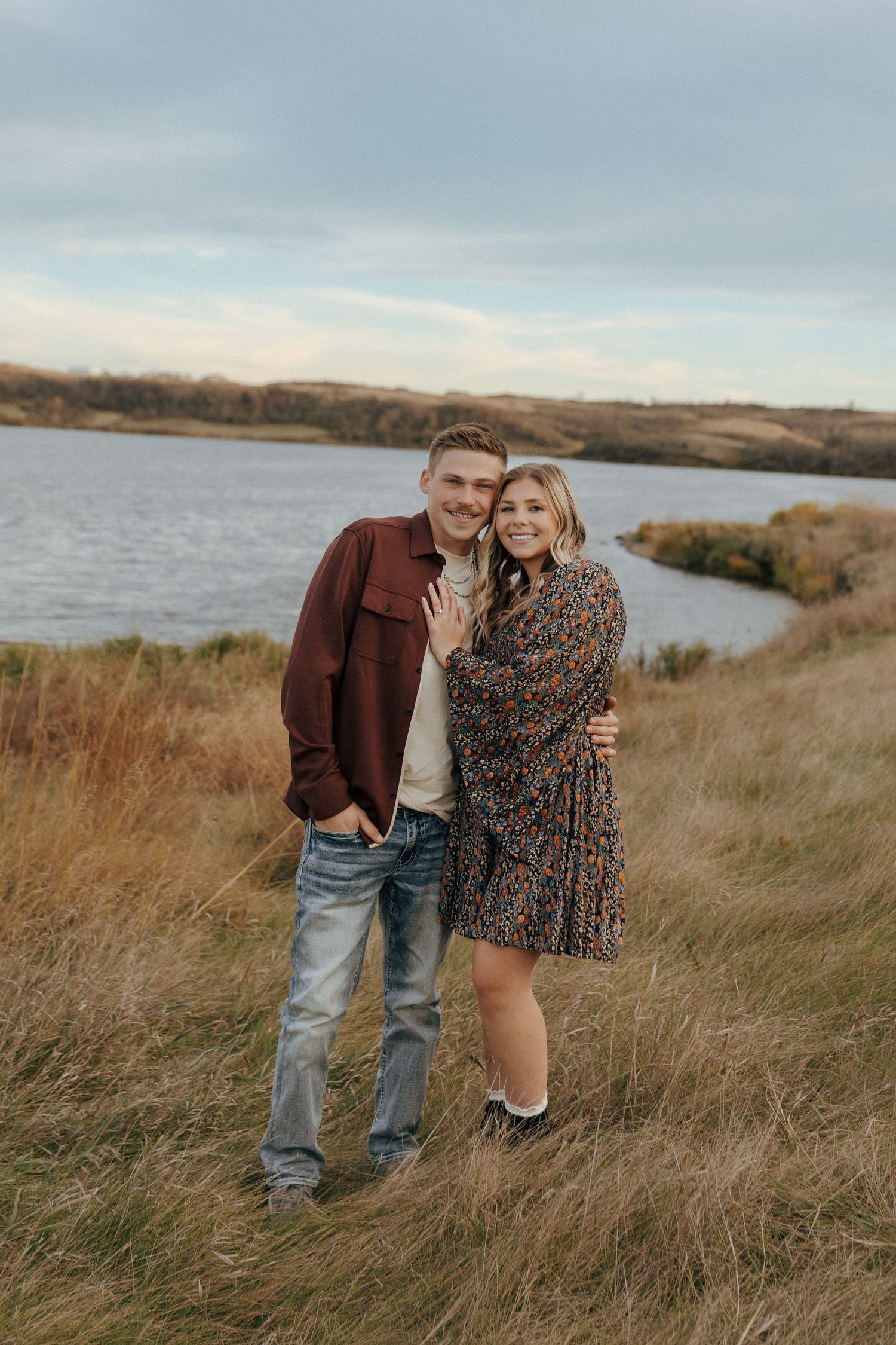 Couple in autumn attire embracing on hillside overlooking lake.