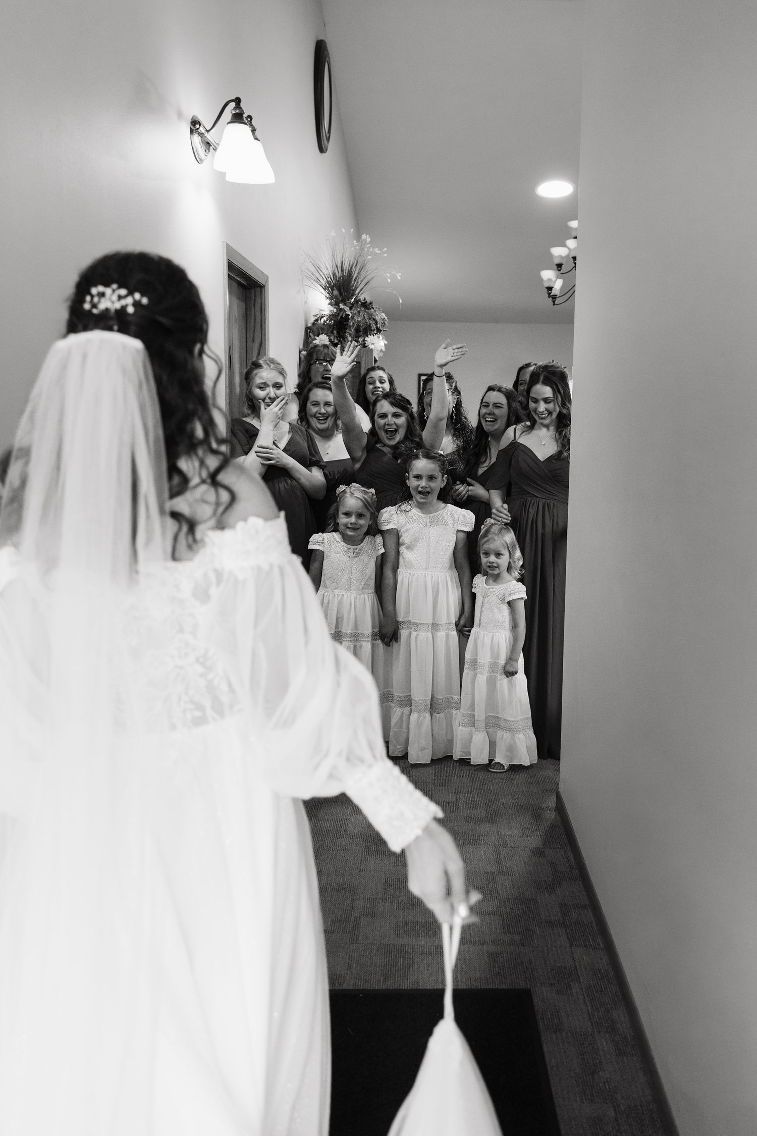 Black and white photo of a bride revealing her wedding dress to excited bridesmaids and flower girls in a hallway.