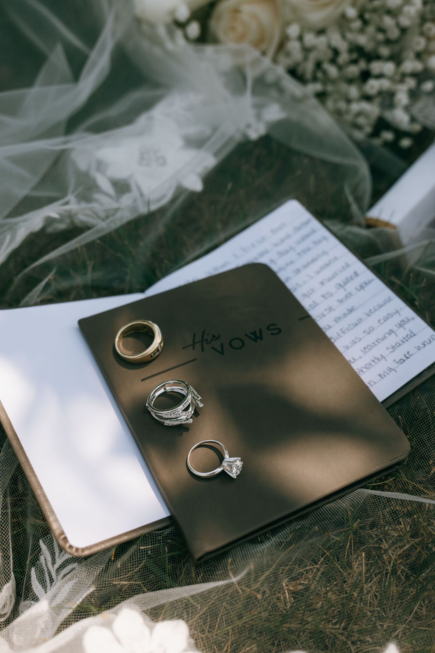 Wedding rings and vow books arranged on grass with bridal veil and flowers in soft natural light.