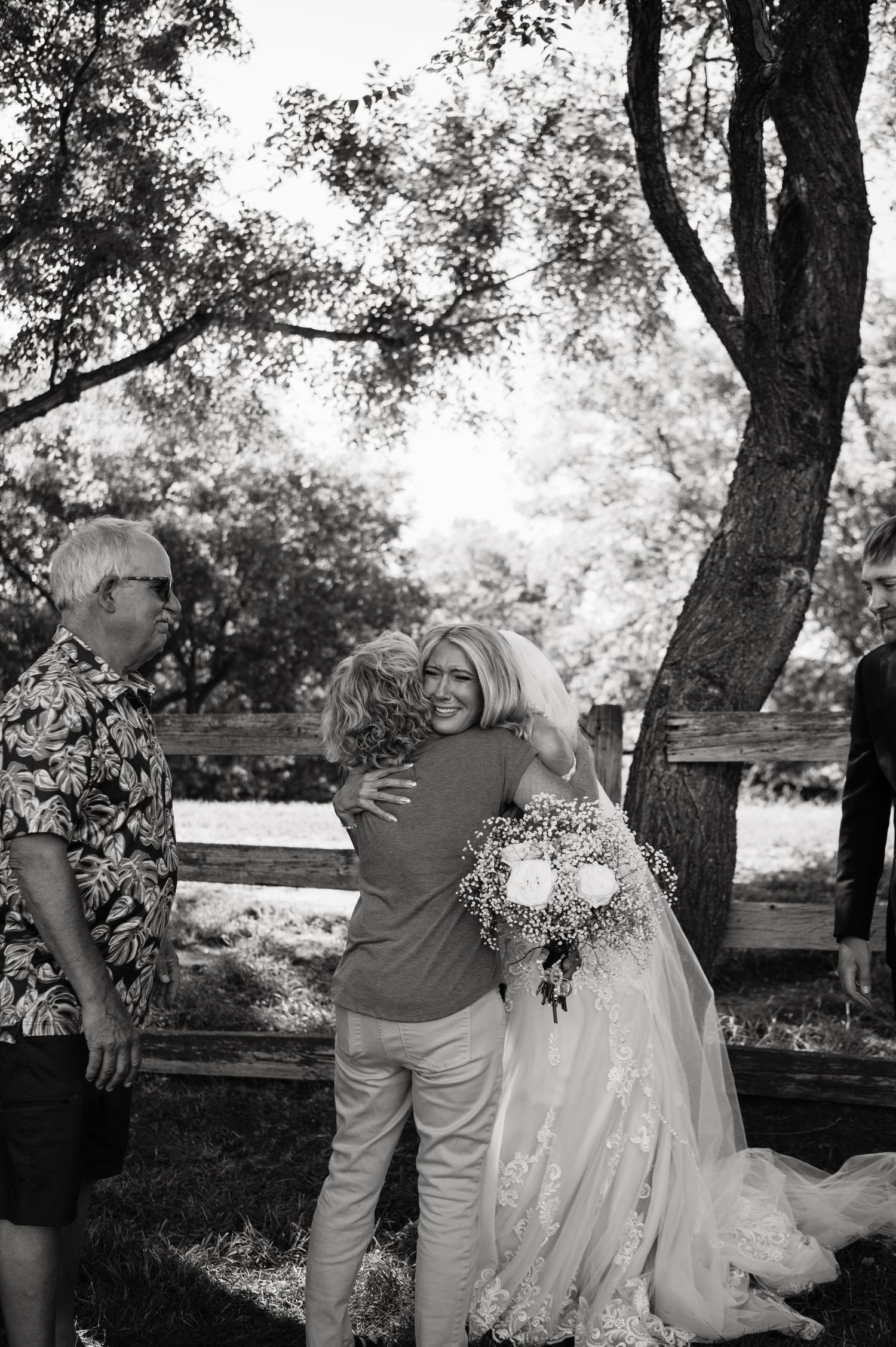 Black and white photo of bride in lace gown embracing guest while holding bouquet at outdoor wedding.