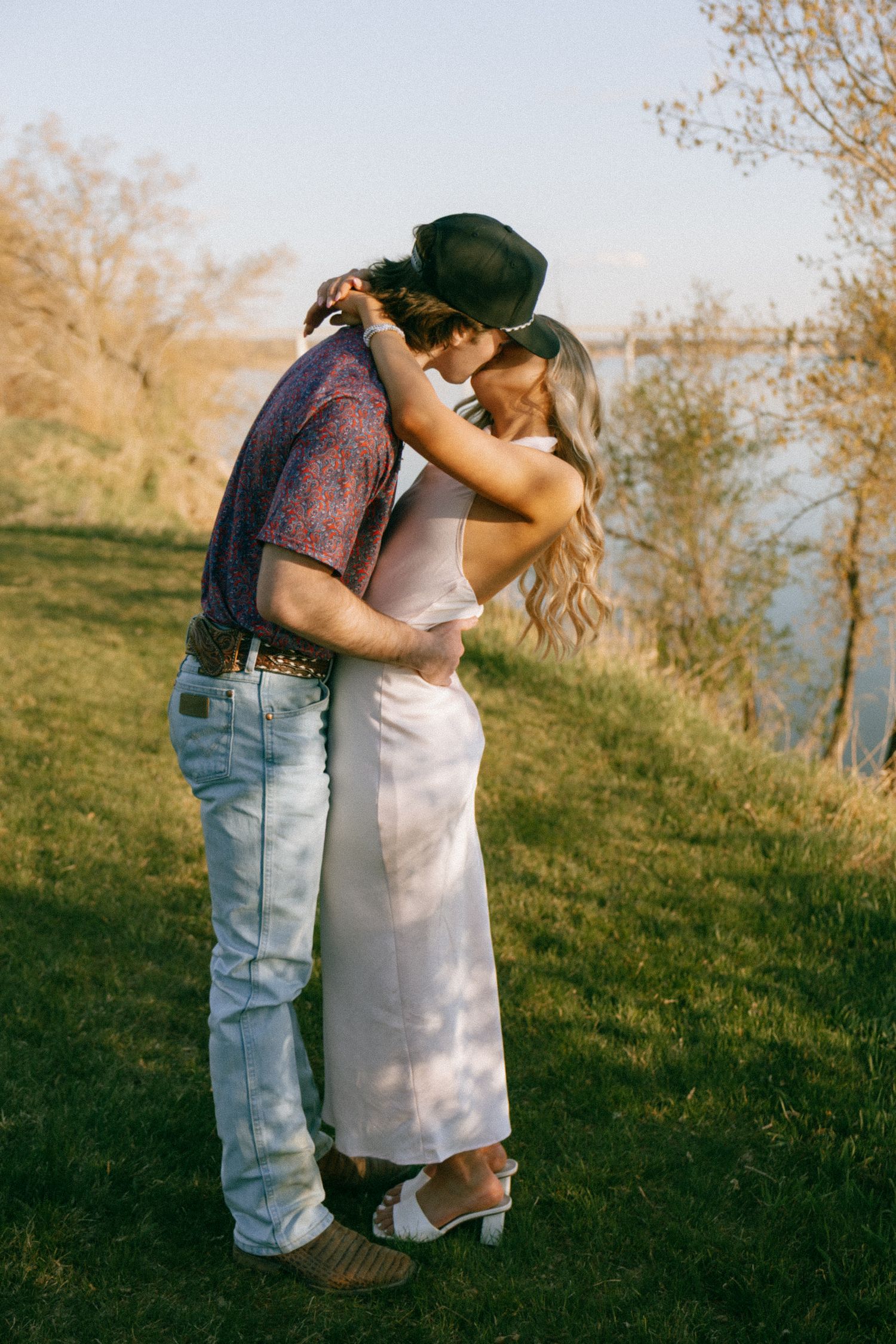 Couple sharing a tender embrace on a grassy riverbank at golden hour in casual summer attire.