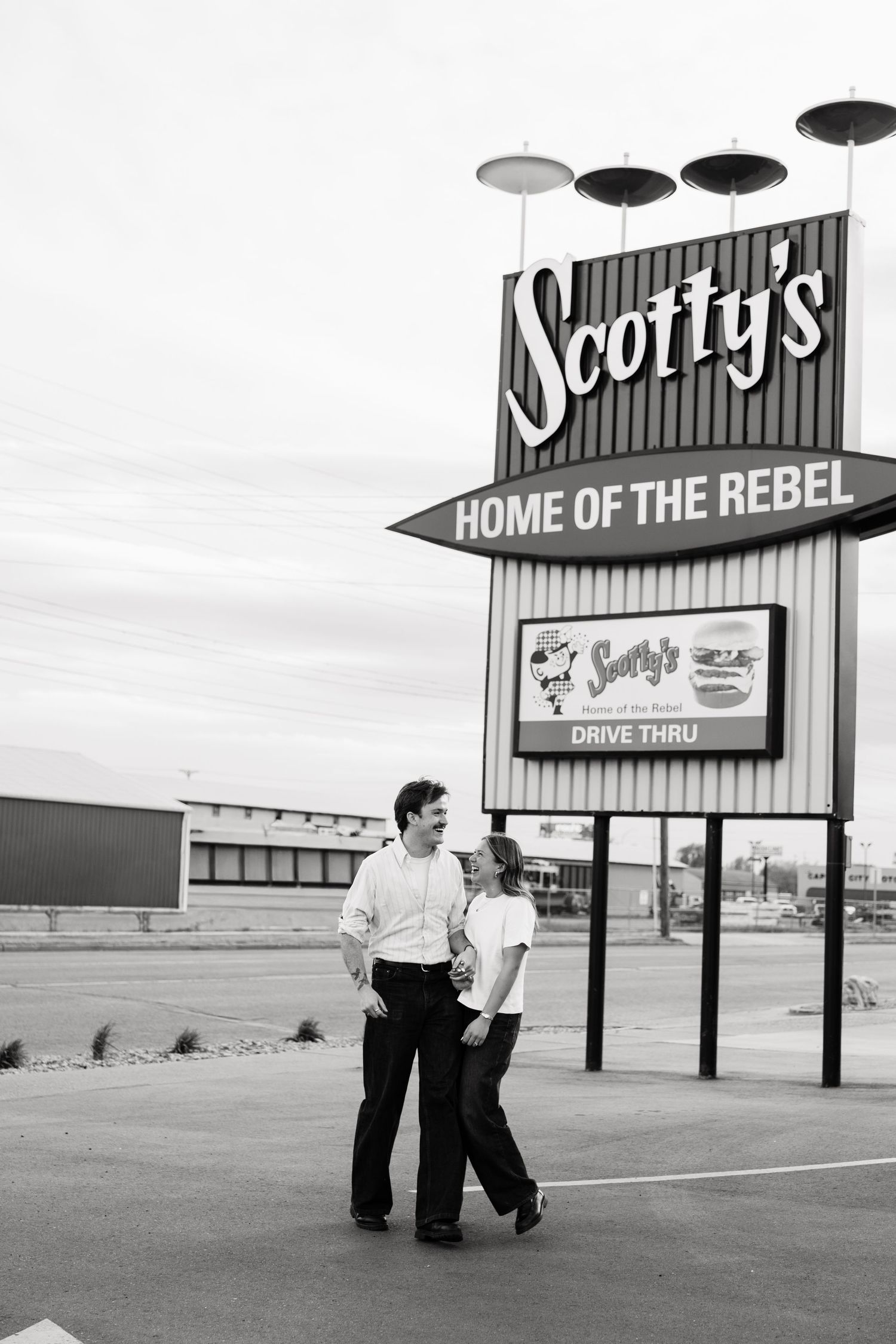 Couple laughing together outside Scotty's Home of the Rebel drive-thru restaurant sign in black and white.