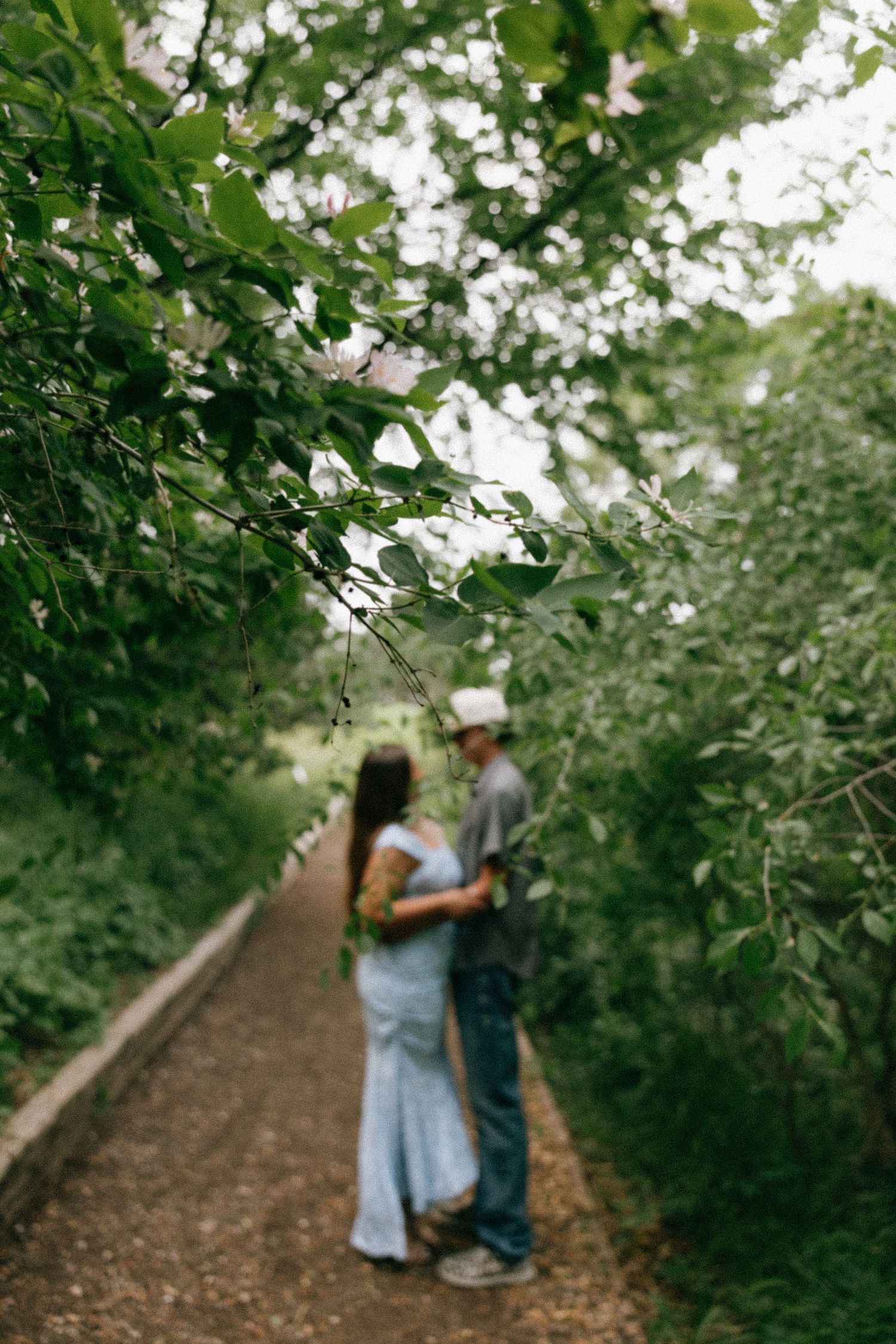 A couple embraces on a lush green tree-lined pathway, woman in blue dress and man in gray shirt and white cap.