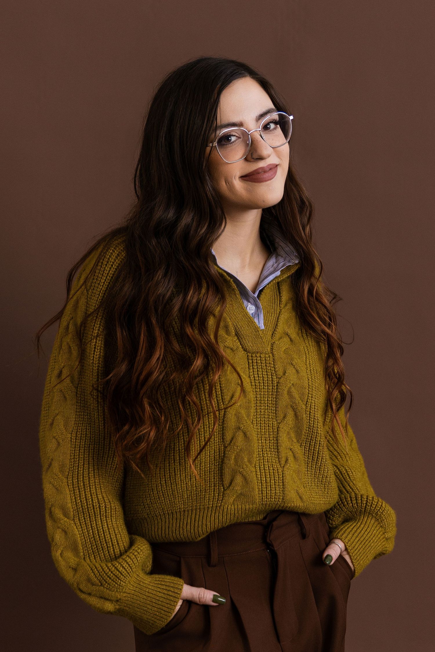 Young woman with long wavy hair wearing olive cable-knit sweater and clear glasses, posing against brown background.