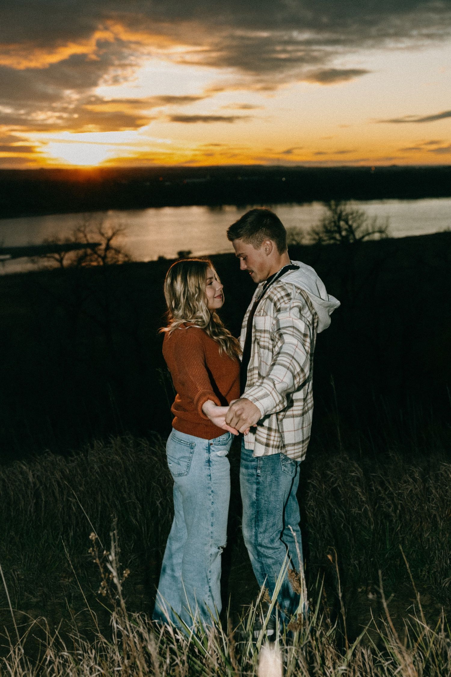 A couple embraces in a field at sunset wearing casual denim and plaid clothing.