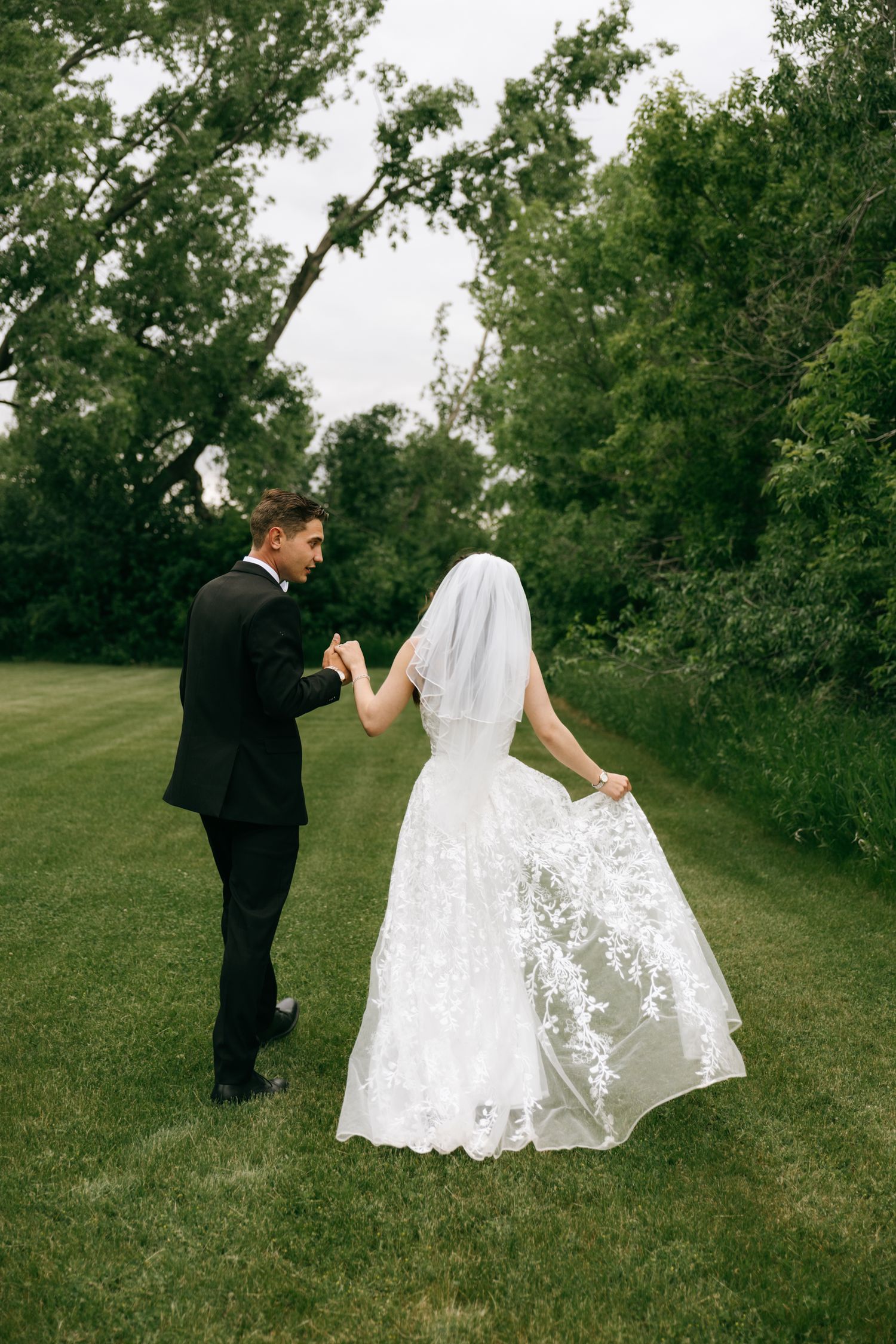 A bride in white lace gown and veil holds hands with groom in black suit while walking on green grass.