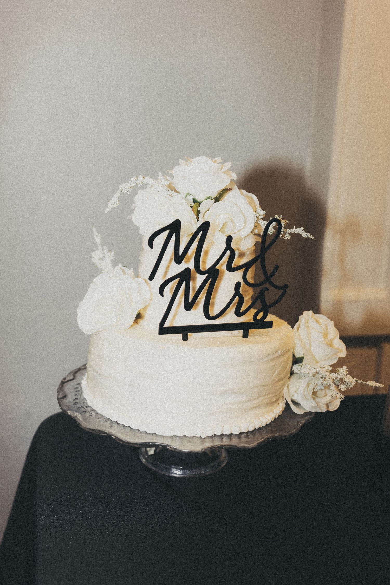 White wedding cake decorated with 'Mr & Mrs' topper and floral accents on dark surface.