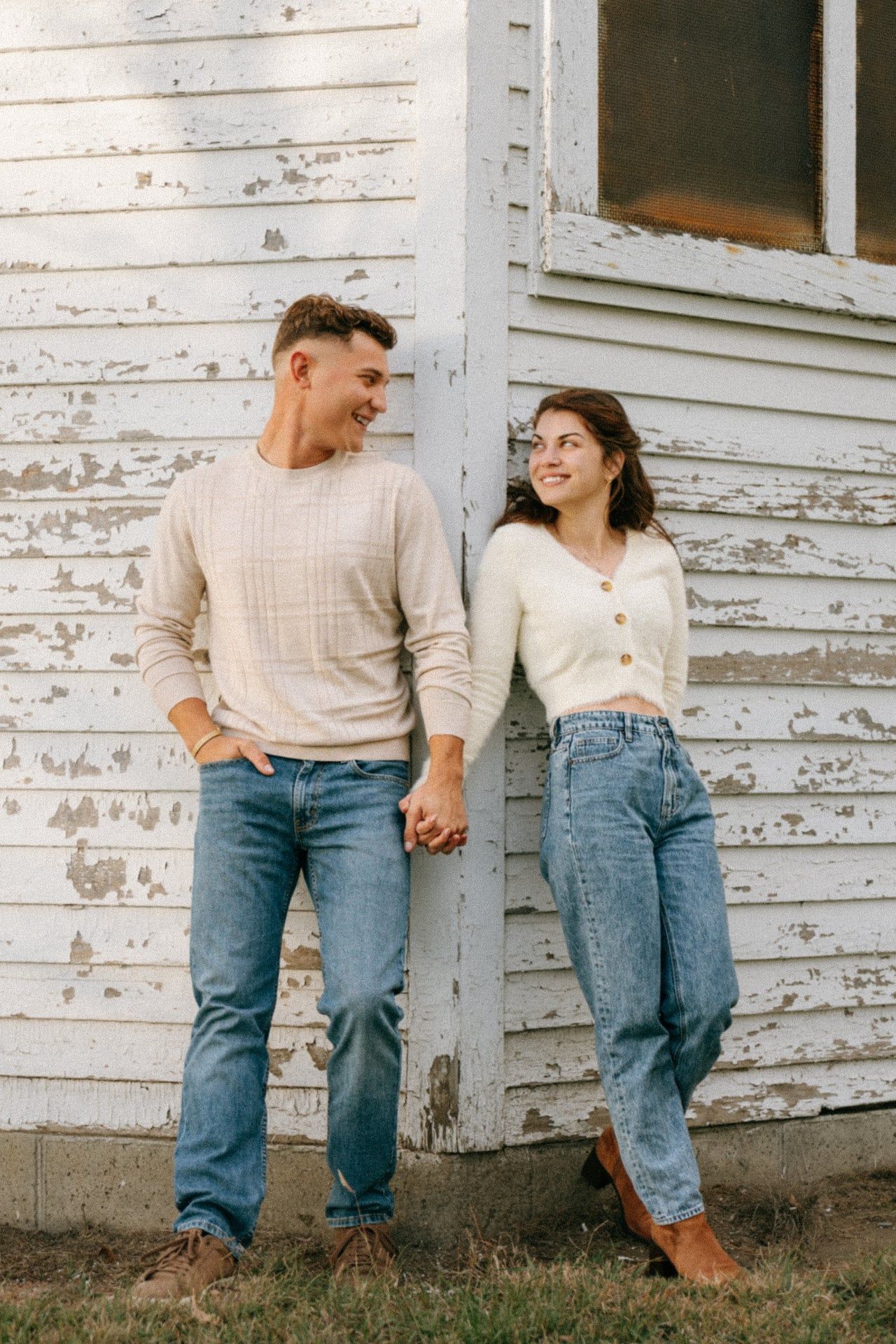 A couple in matching cream sweaters and blue jeans lean against a white weathered wall in a romantic engagement photoshoot in Bismarck.
