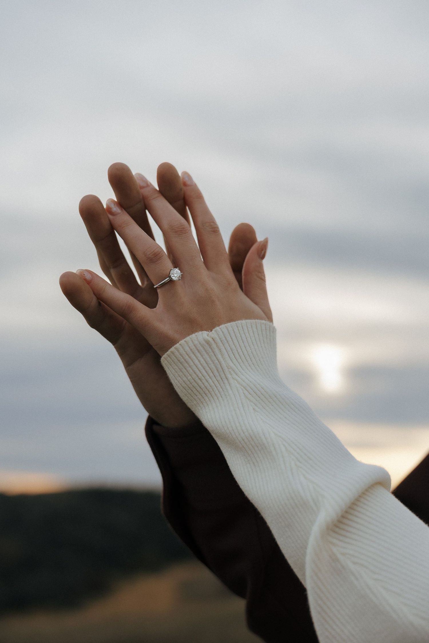 Two hands intertwined against a cloudy sky, showcasing an engagement ring.