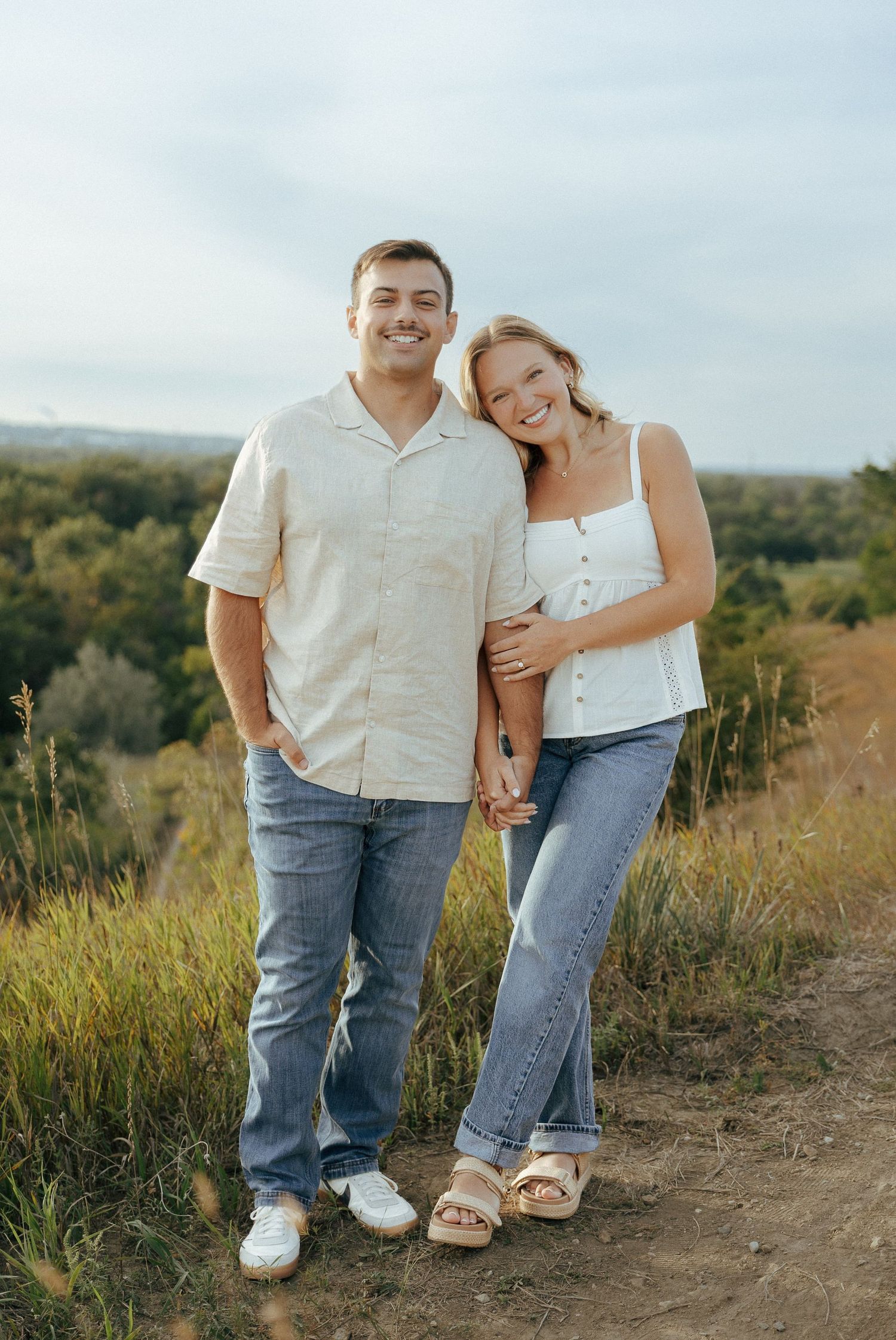 Two people in matching white shirts and jeans stand together on a scenic hiking trail.