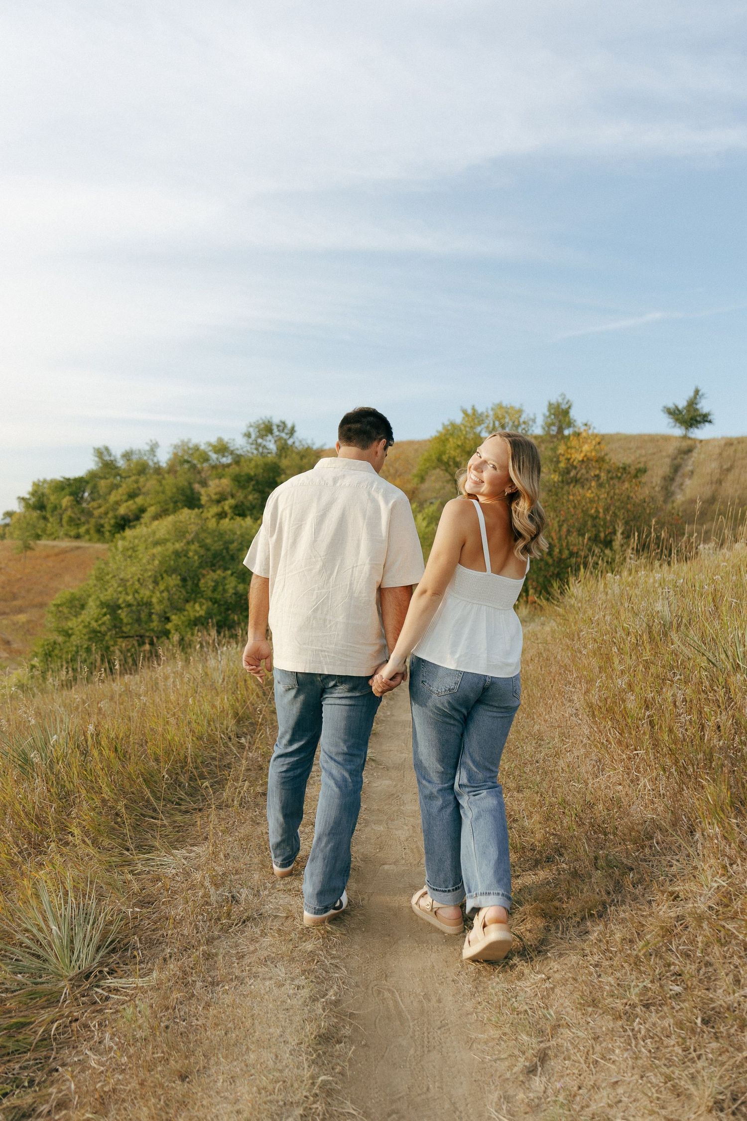 A couple walks together on a scenic hillside path surrounded by dry grass and blue skies.