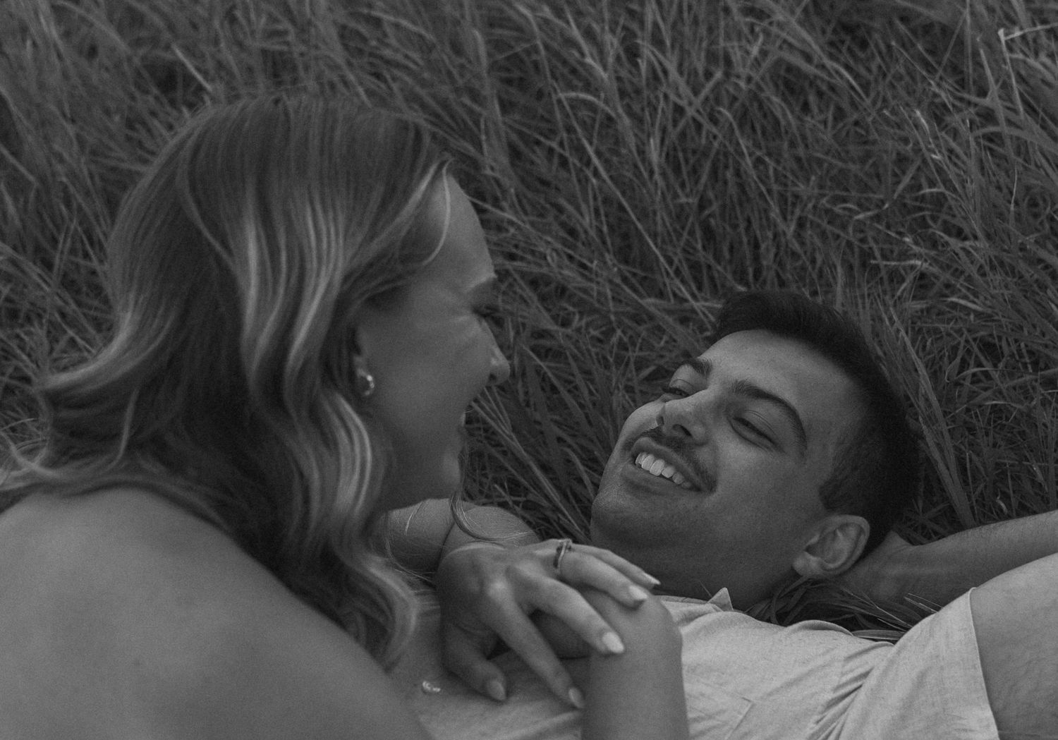 A black and white candid photo of a couple lying in tall grass sharing a laugh.