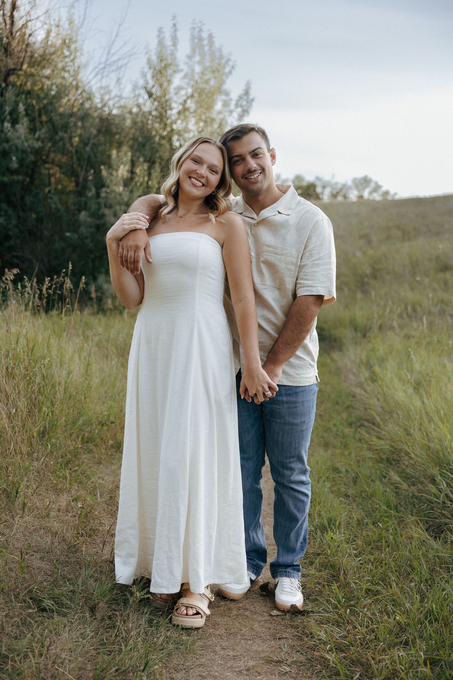 A couple in coordinating white and casual outfits pose together on a nature trail.