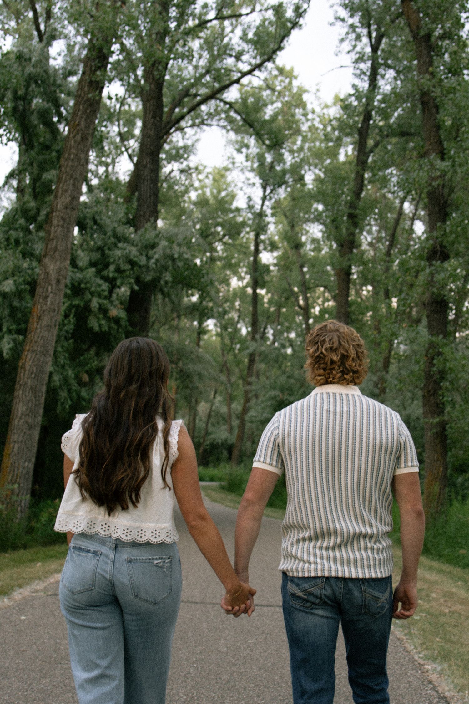 Couple holding hands walking down a tree-lined path, viewed from behind, wearing casual jeans and tops.