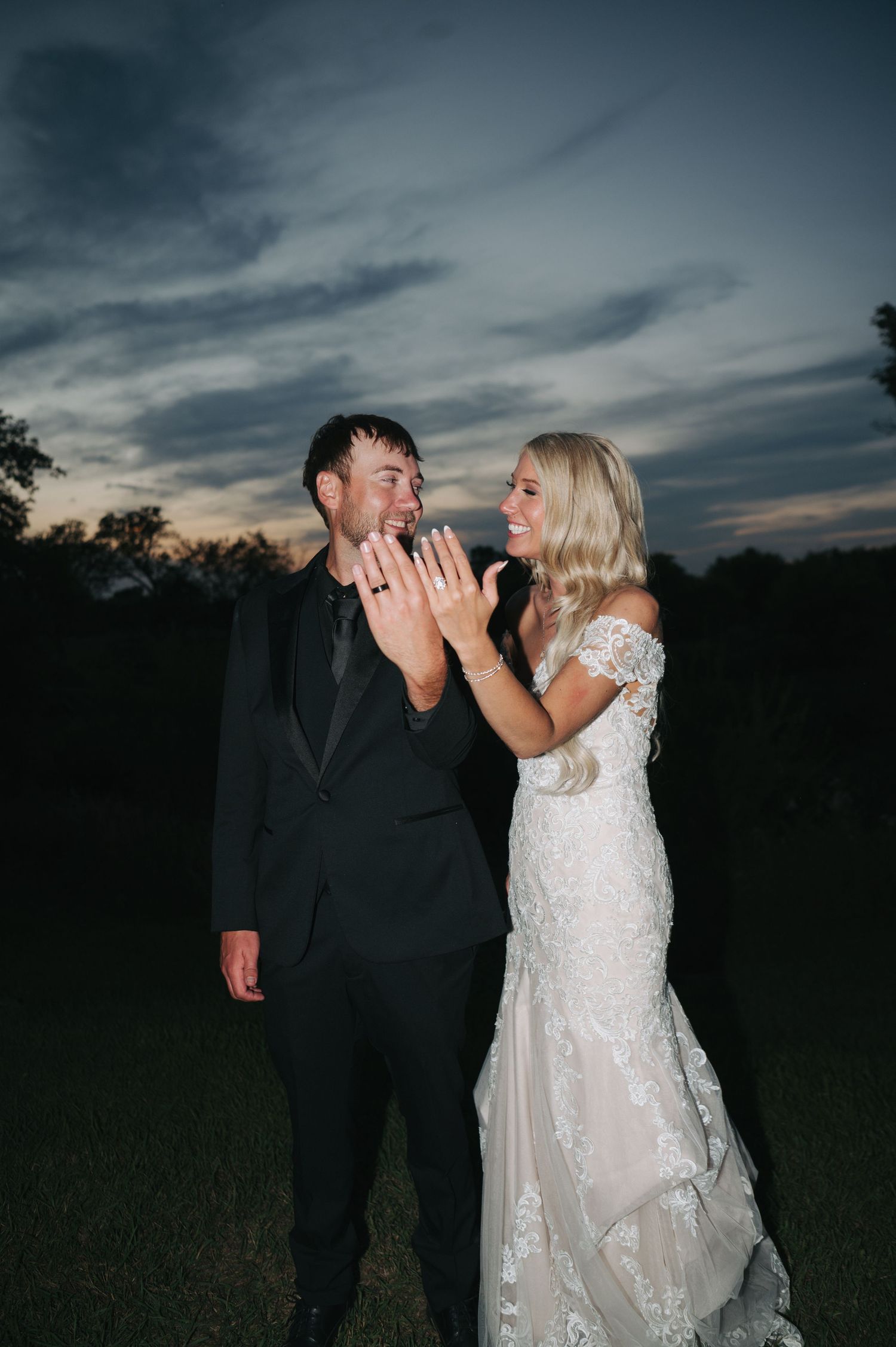 A bride and groom smile while showing off their wedding rings outdoors at sunset, she in lace gown, he in black tux.