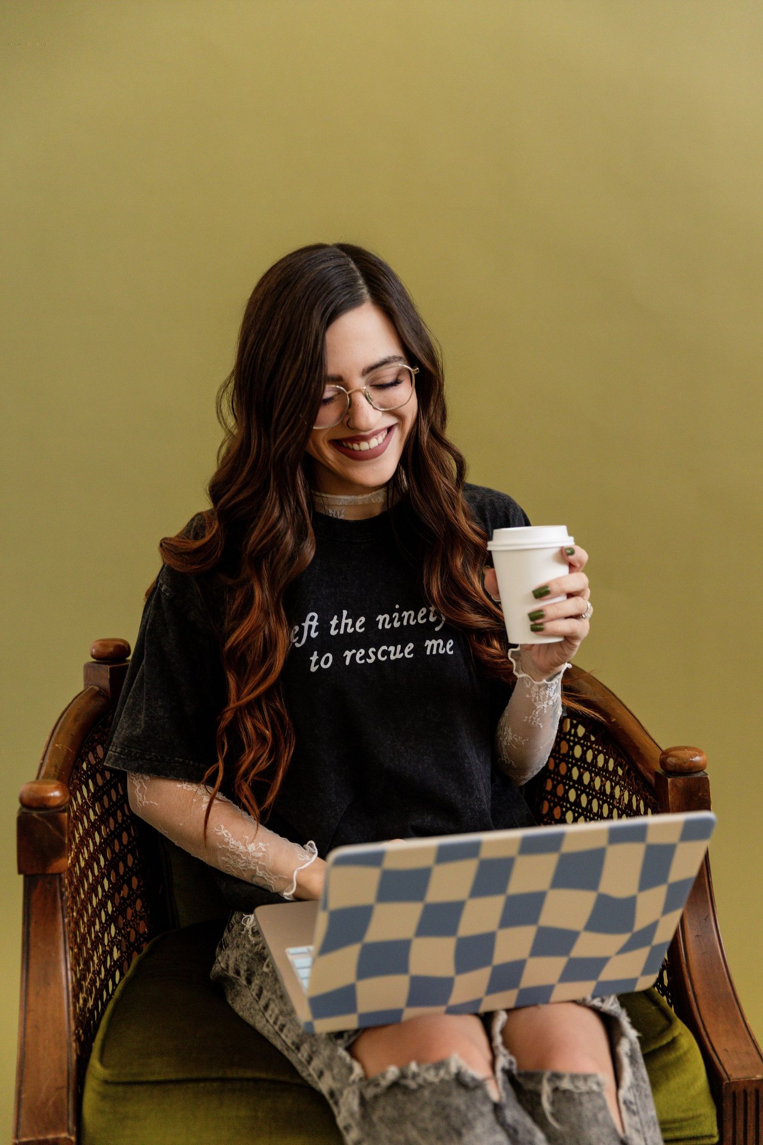 Smiling woman with long brown hair holding coffee cup, using checkered laptop while sitting in vintage chair.