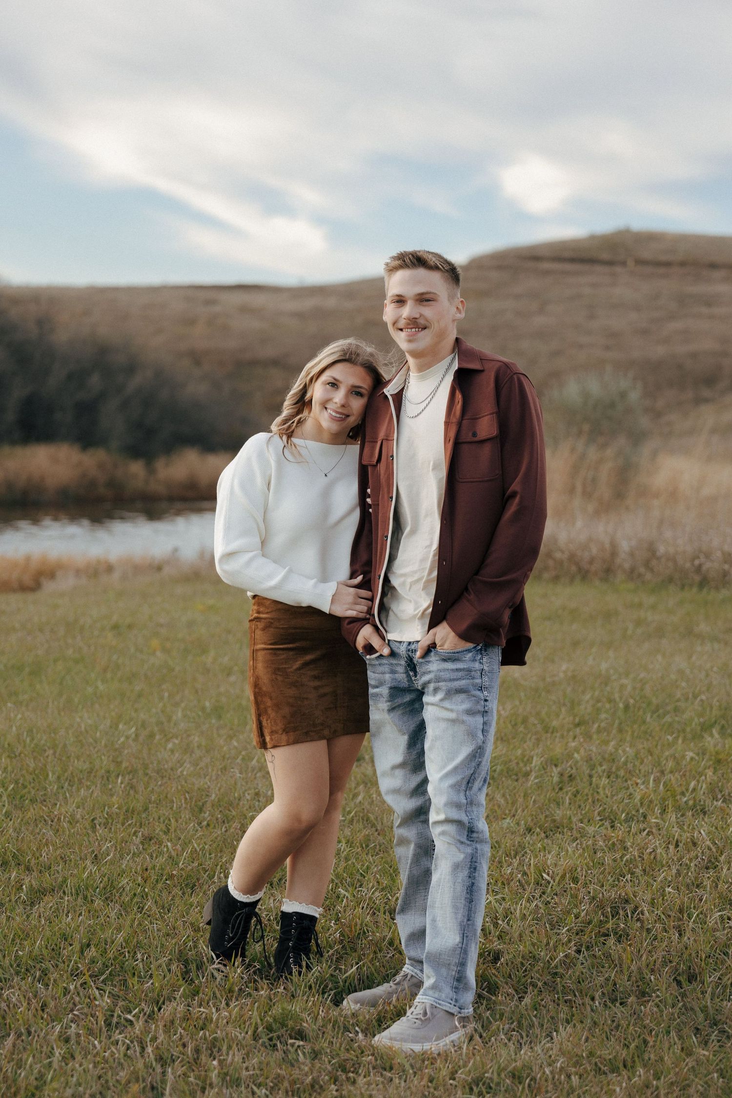 Two people share an intimate moment in a scenic field with rolling hills and autumn foliage.