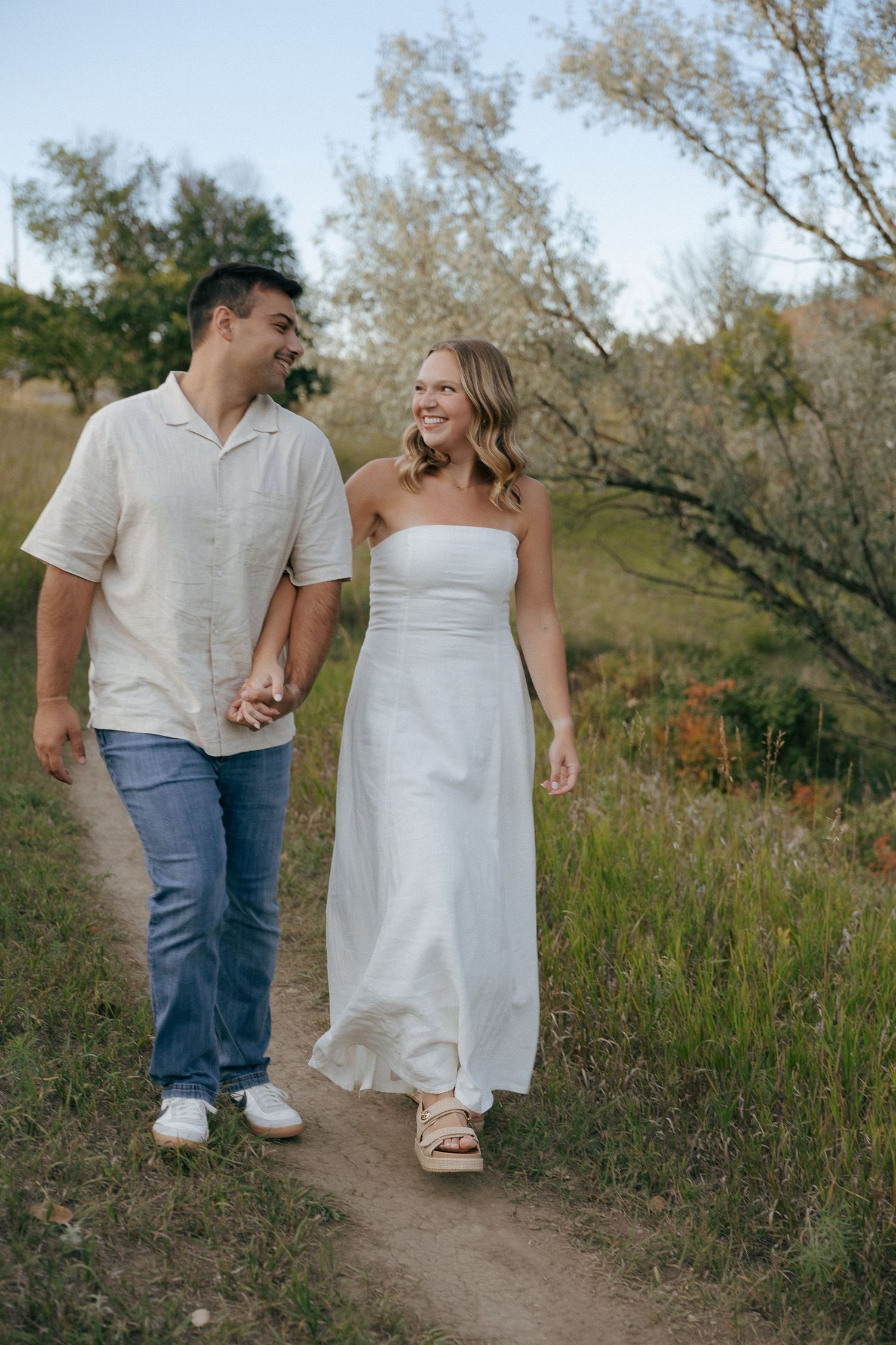 A couple walks hand in hand along a dirt path surrounded by natural scenery.