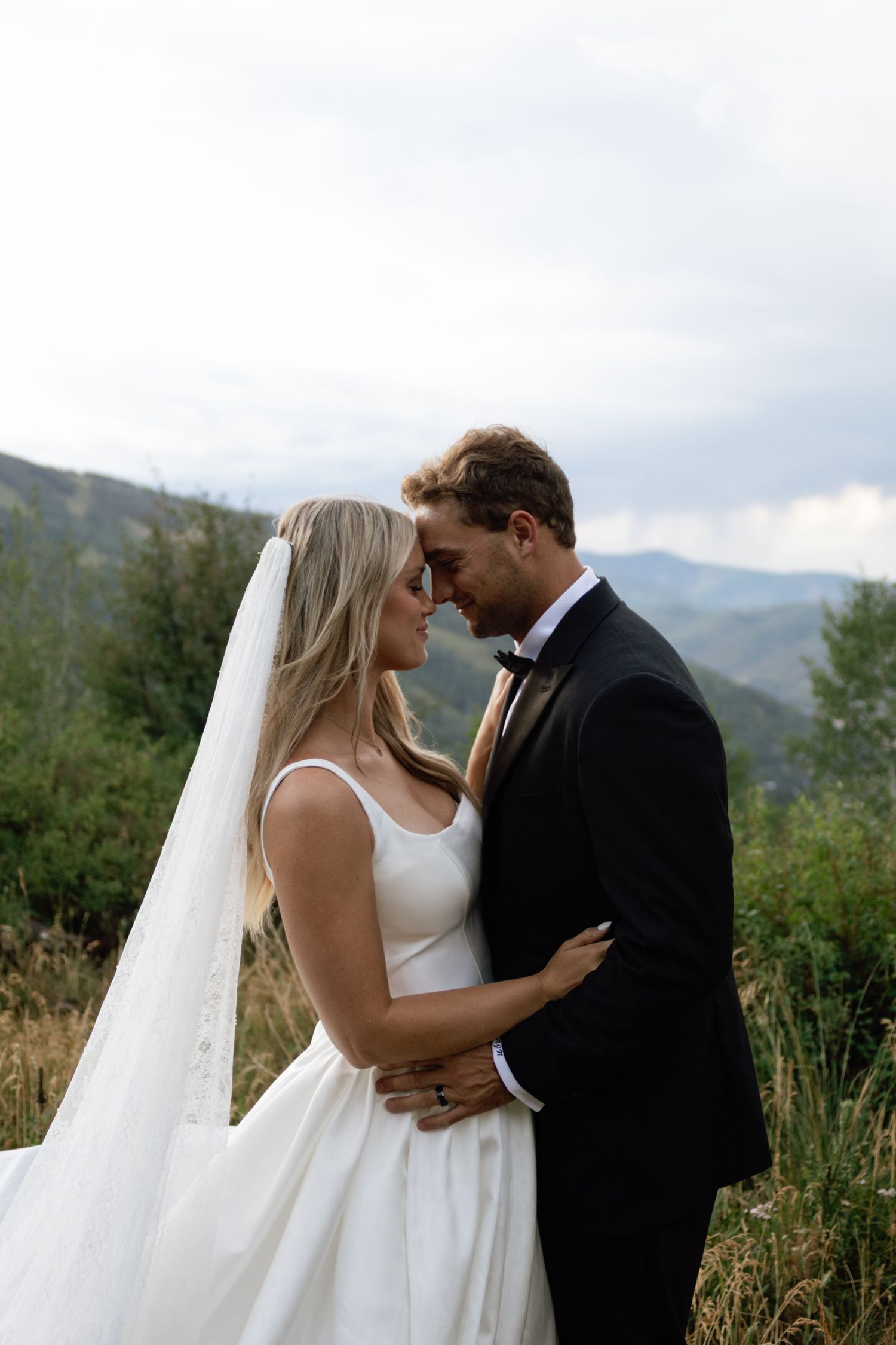 Bride and groom embrace forehead-to-forehead in white gown and black tuxedo with mountain landscape backdrop.