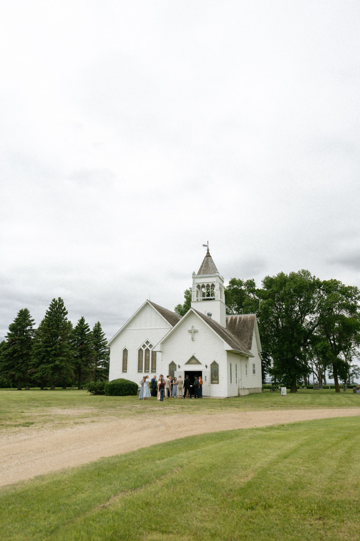 A white country church with a cross steeple surrounded by green trees, with wedding guests gathered at the entrance on a cloudy day.