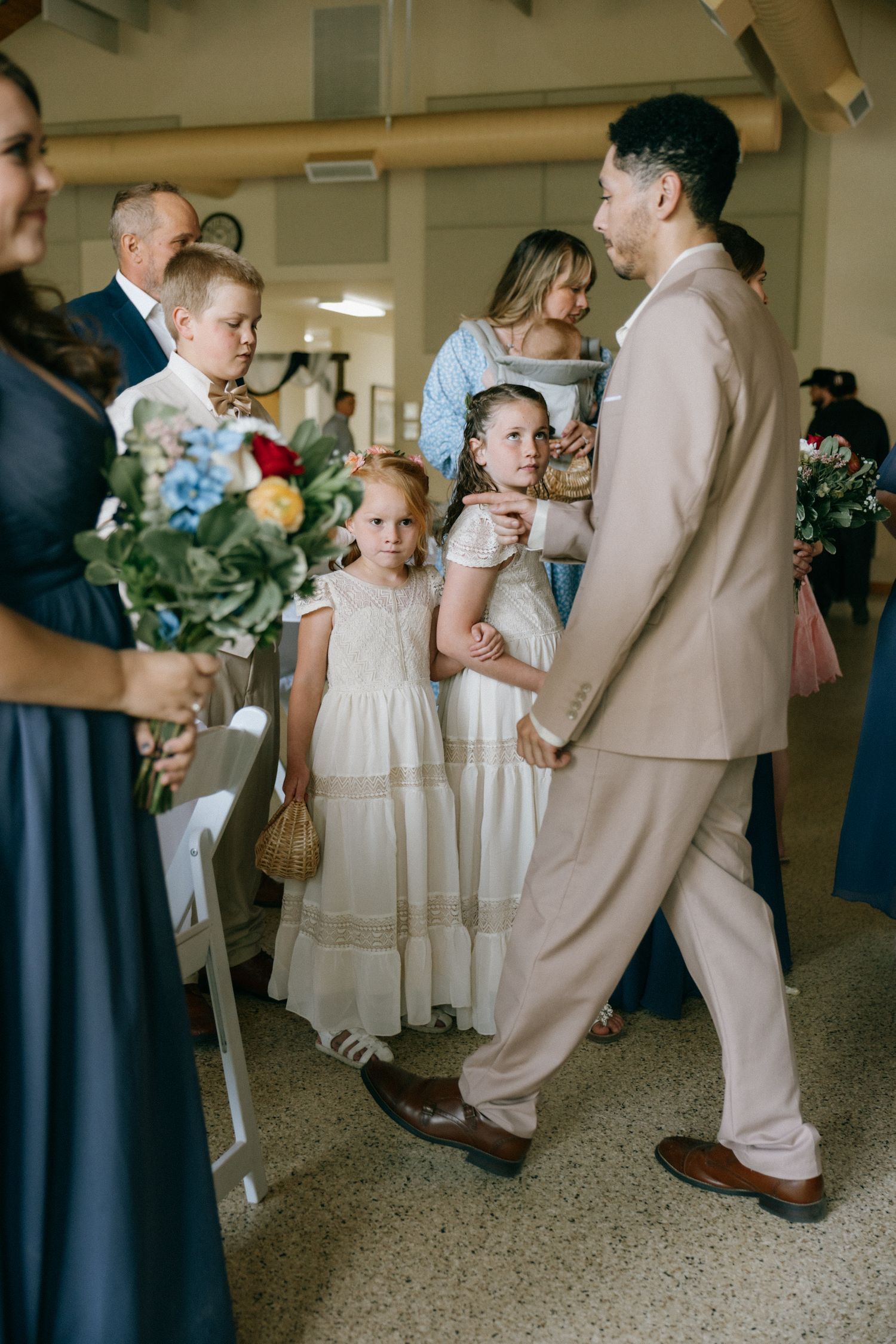 Flower girls in white dresses walk with a man in a beige suit during a wedding ceremony indoors.
