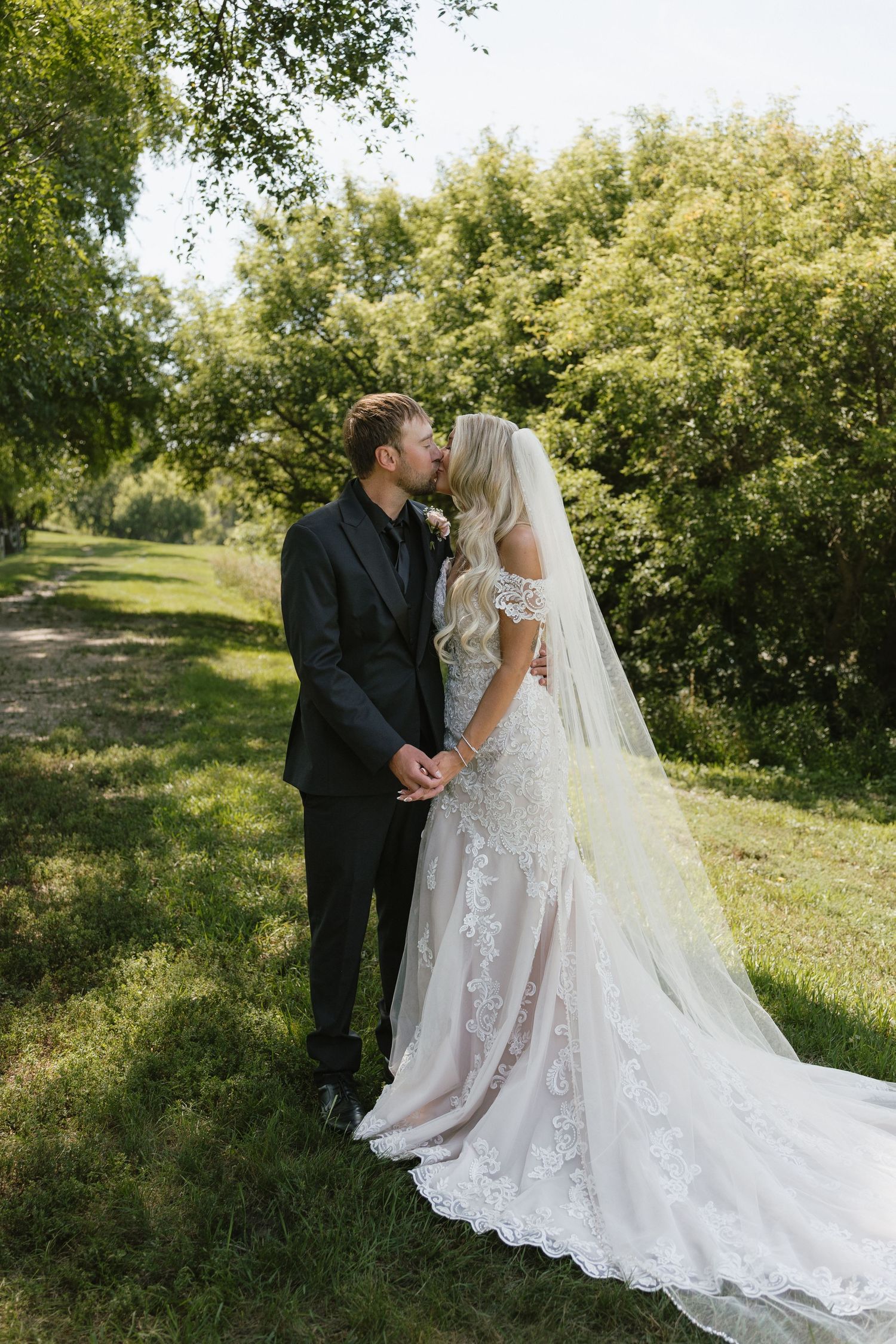 Bride and groom sharing a kiss on grass path, bride in lace gown with flowing veil, groom in black suit.