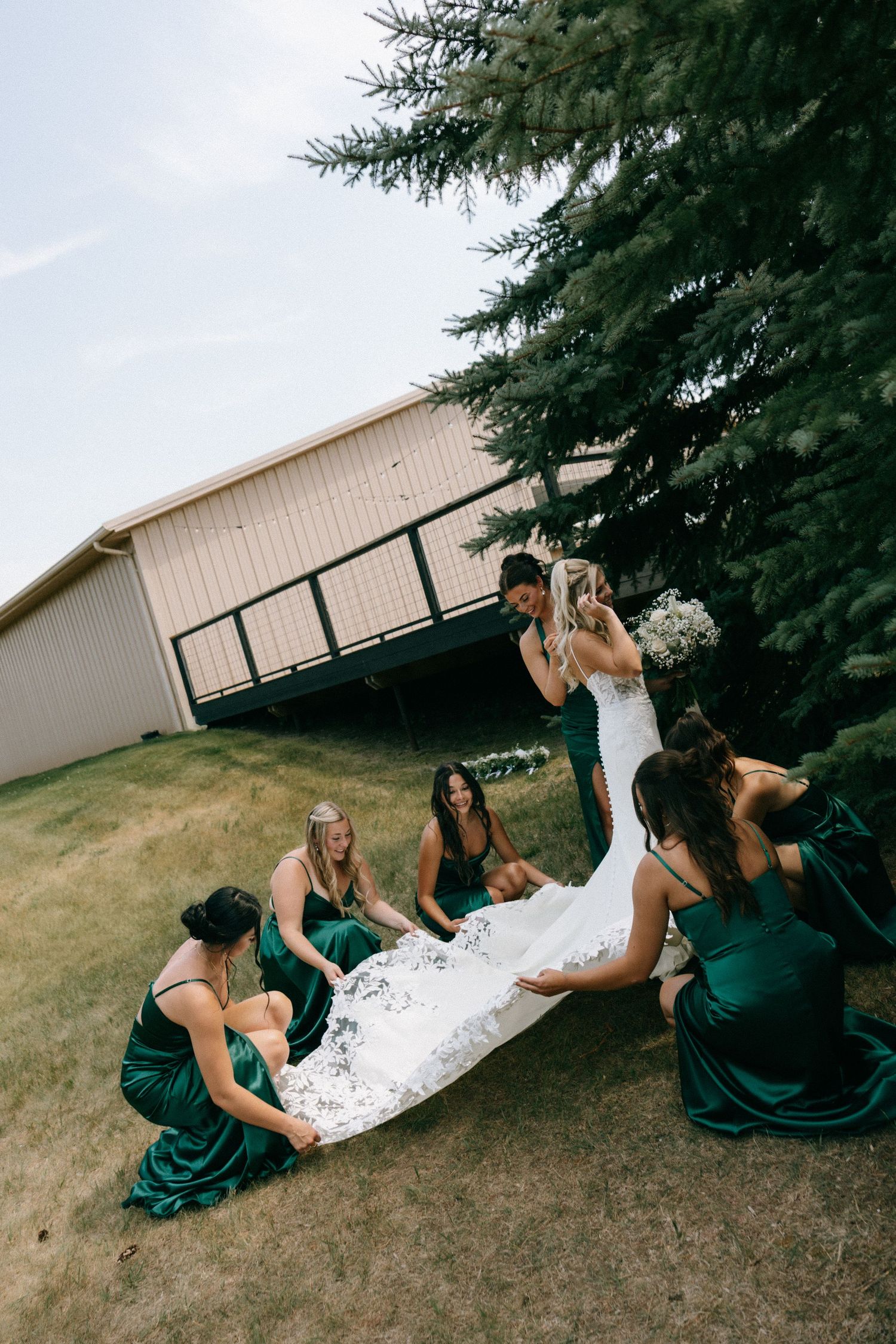 Bridesmaids in emerald green dresses arrange the bride's white lace wedding gown train outdoors near a pine tree.
