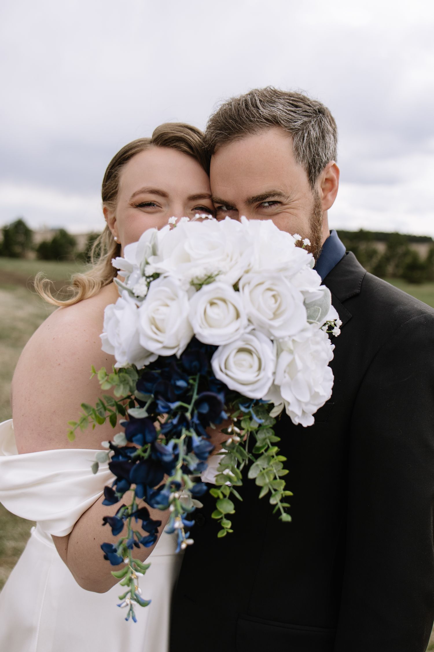 Bride and groom smiling behind a white rose and blue flower bouquet outdoors on their wedding day.