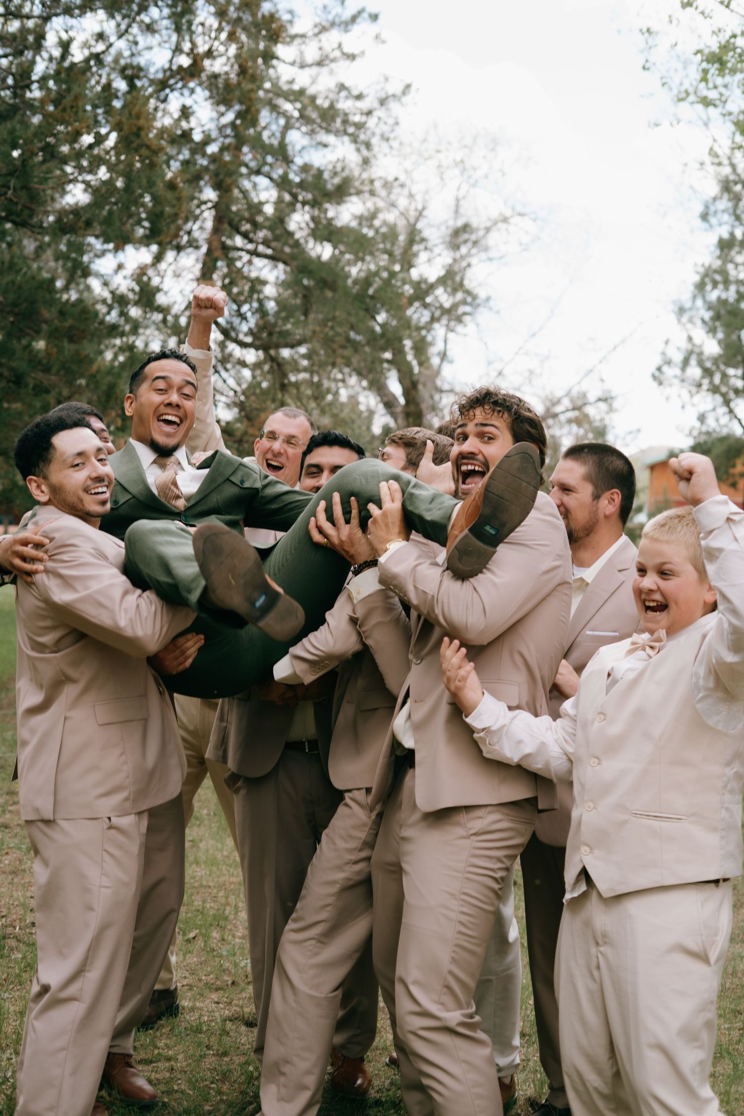 Groomsmen in tan suits joyfully lifting the groom outdoors at a wedding celebration.