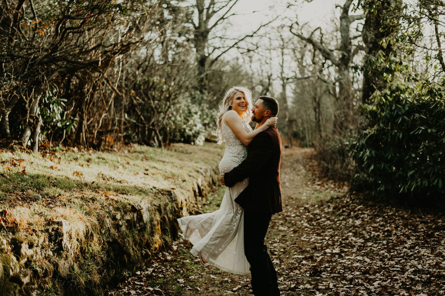 A couple shares an intimate moment on their wedding day in a sunlit wooded area with autumn leaves on the ground.