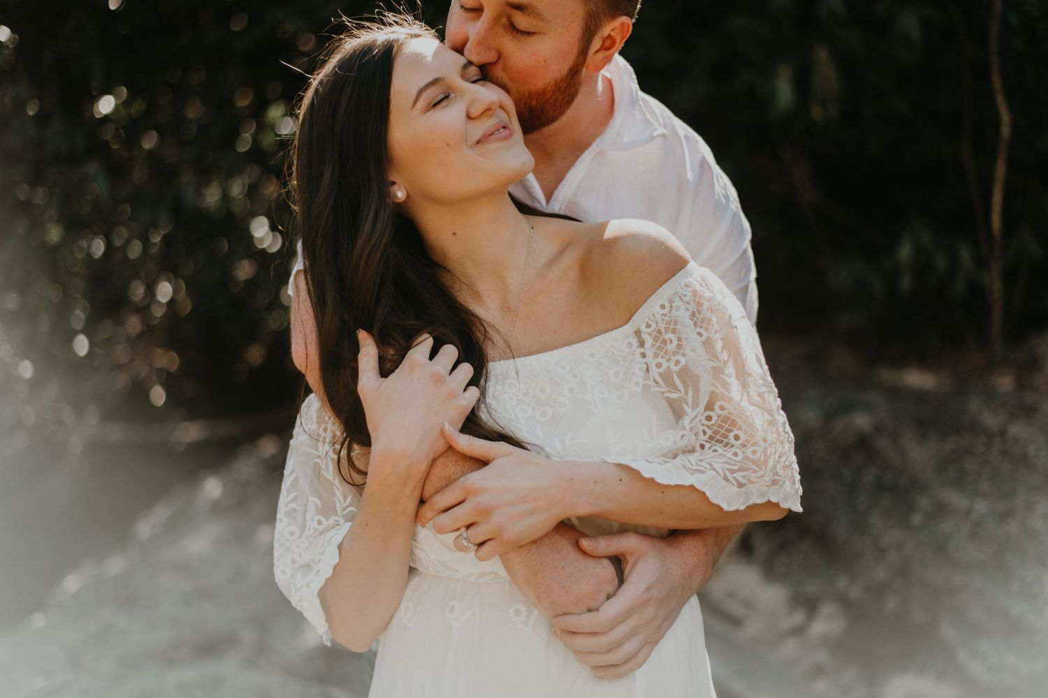 An intimate embrace between a couple in wedding attire against a sunlit natural background.