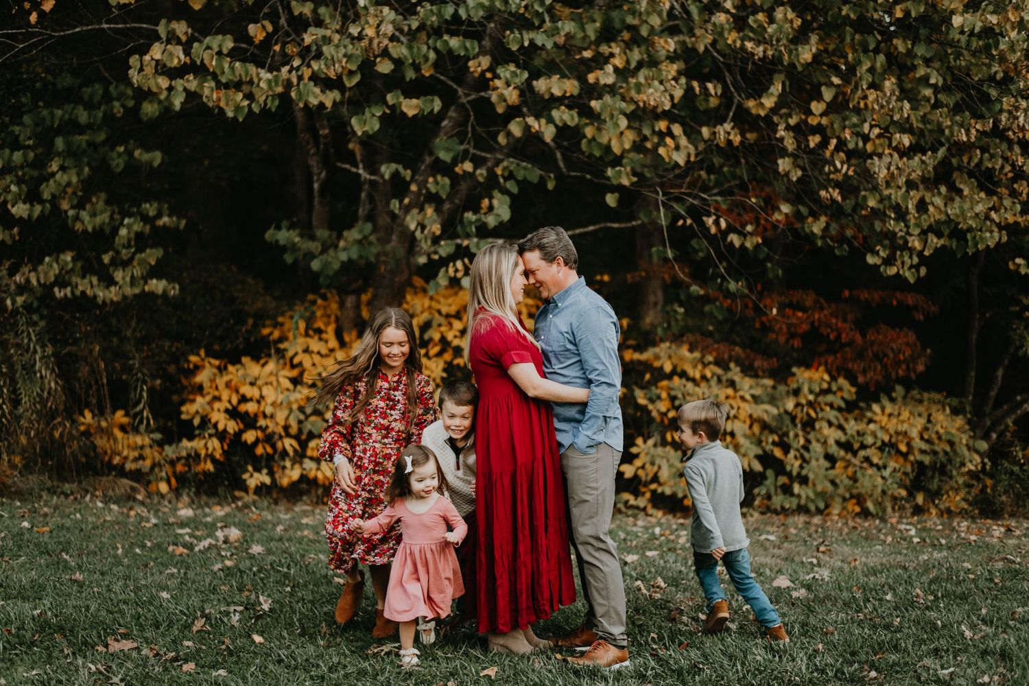 A family wearing red and blue clothing poses together during a fall photoshoot in a park with autumn foliage.