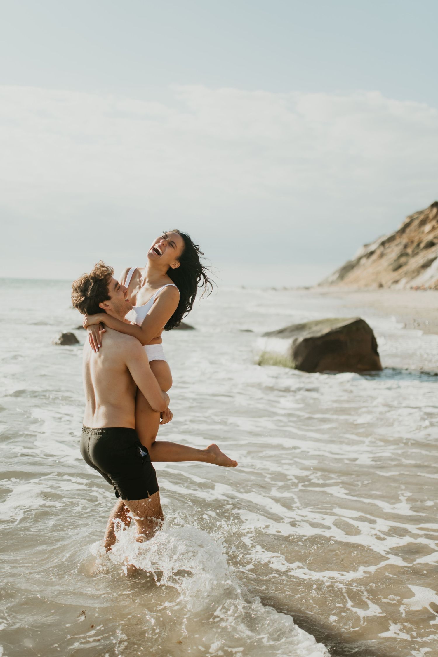 Two figures splash and play together in ocean waves near a sandy beach during a sunny day.