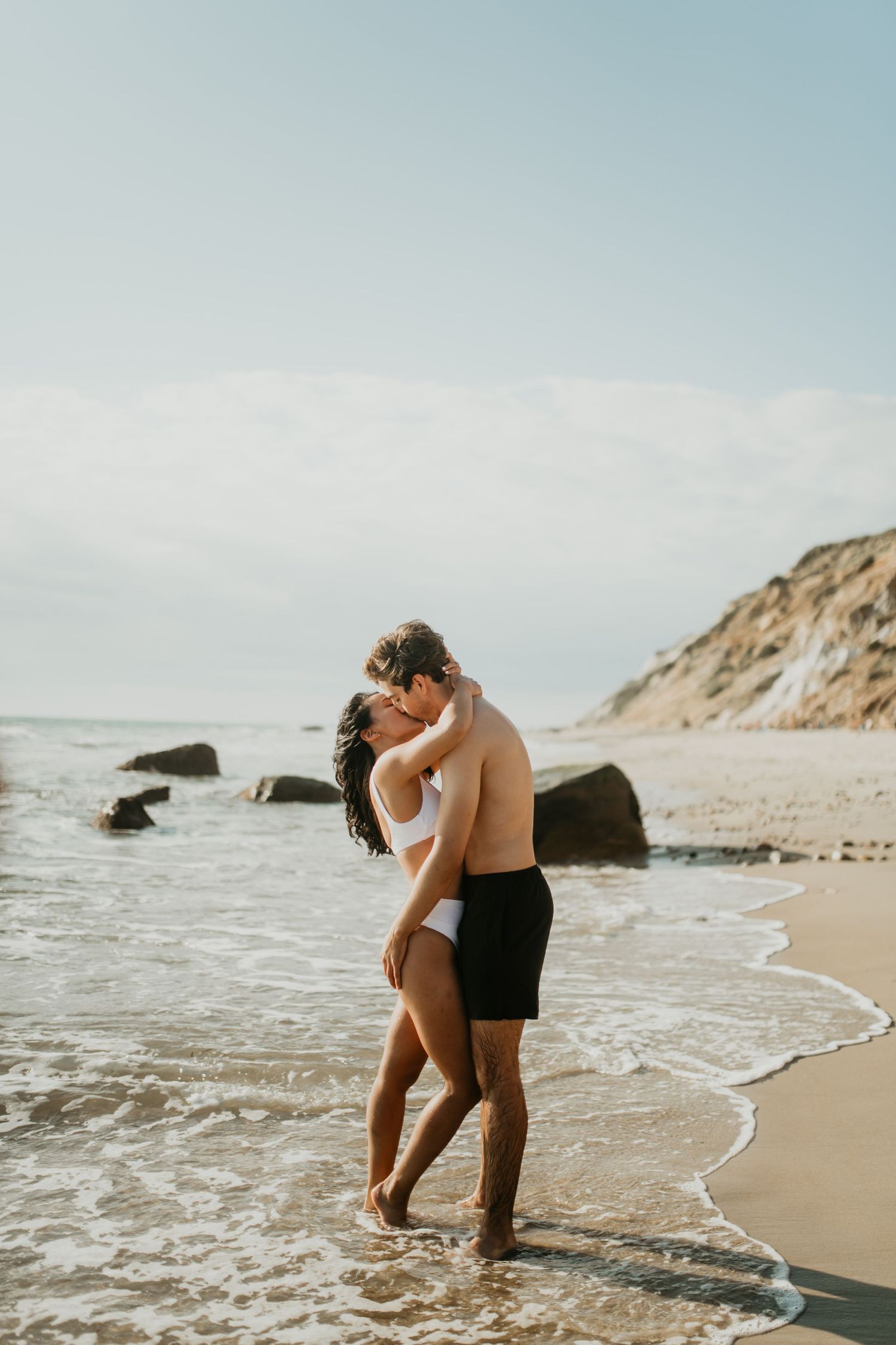 A couple embraces on a scenic beach with cliffs and waves at sunset creating a romantic coastal moment.