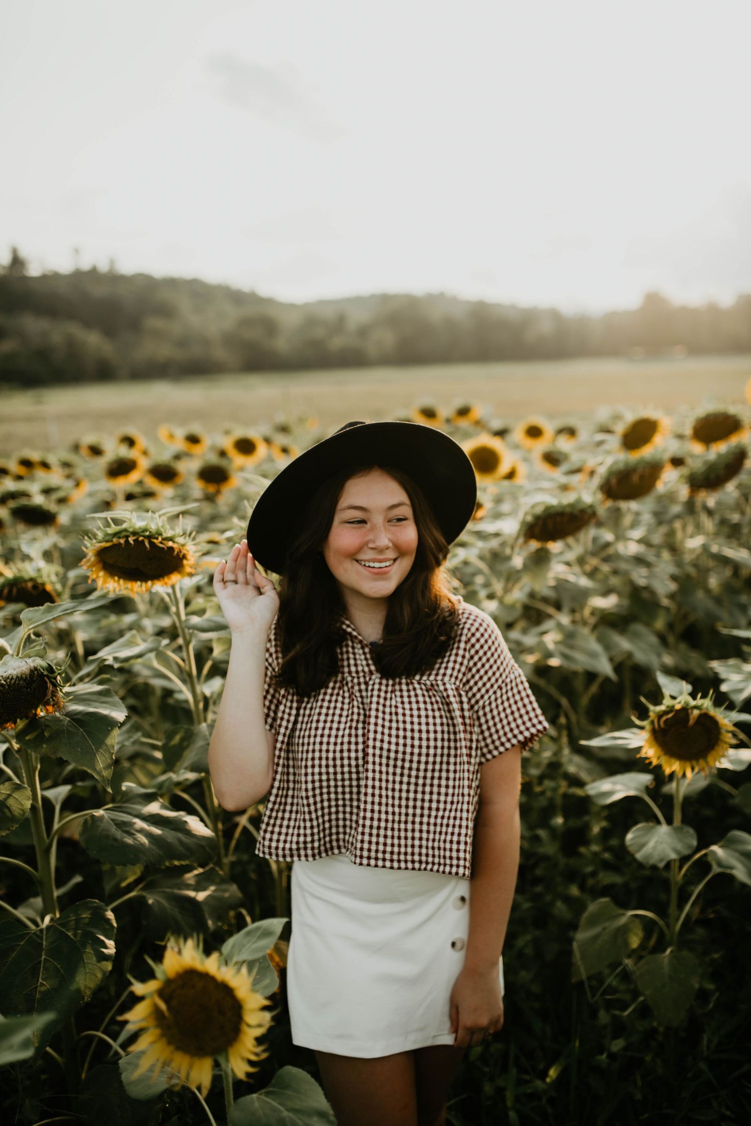 A sun-drenched sunflower field glows at golden hour as someone in a wide-brimmed hat and checkered top poses amid the blooms.