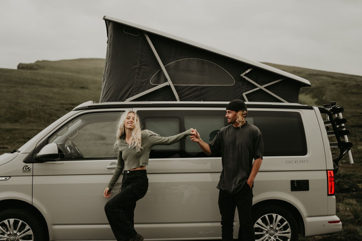 Two people stand beside a silver camper van with a pop-up roof on a cloudy hillside.