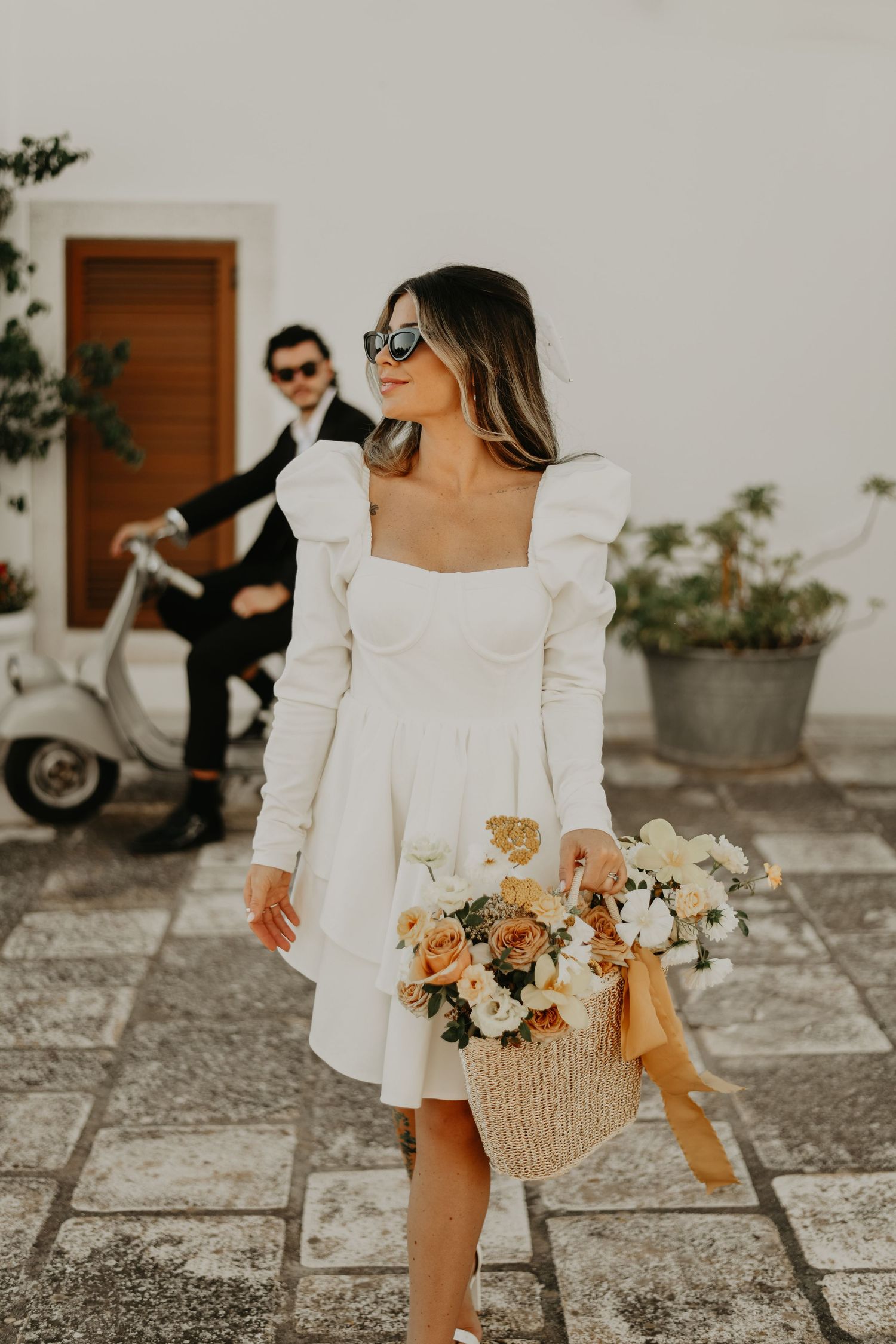 Bride in modern white dress holding floral basket with vintage Vespa in background in Puglia Italy.