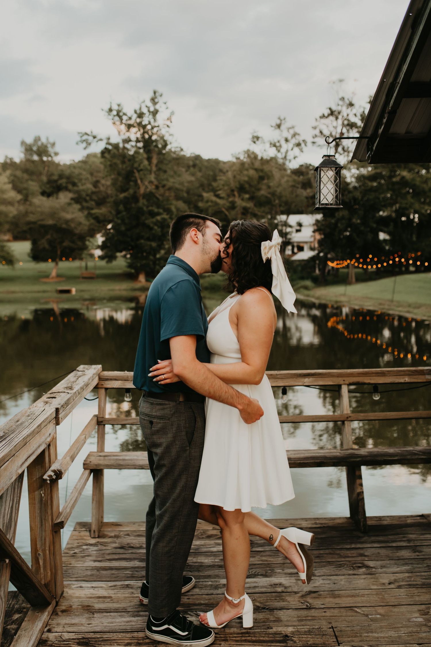 A romantic couple shares intimate moments on a wooden dock by a lake at sunset with string lights in the background.